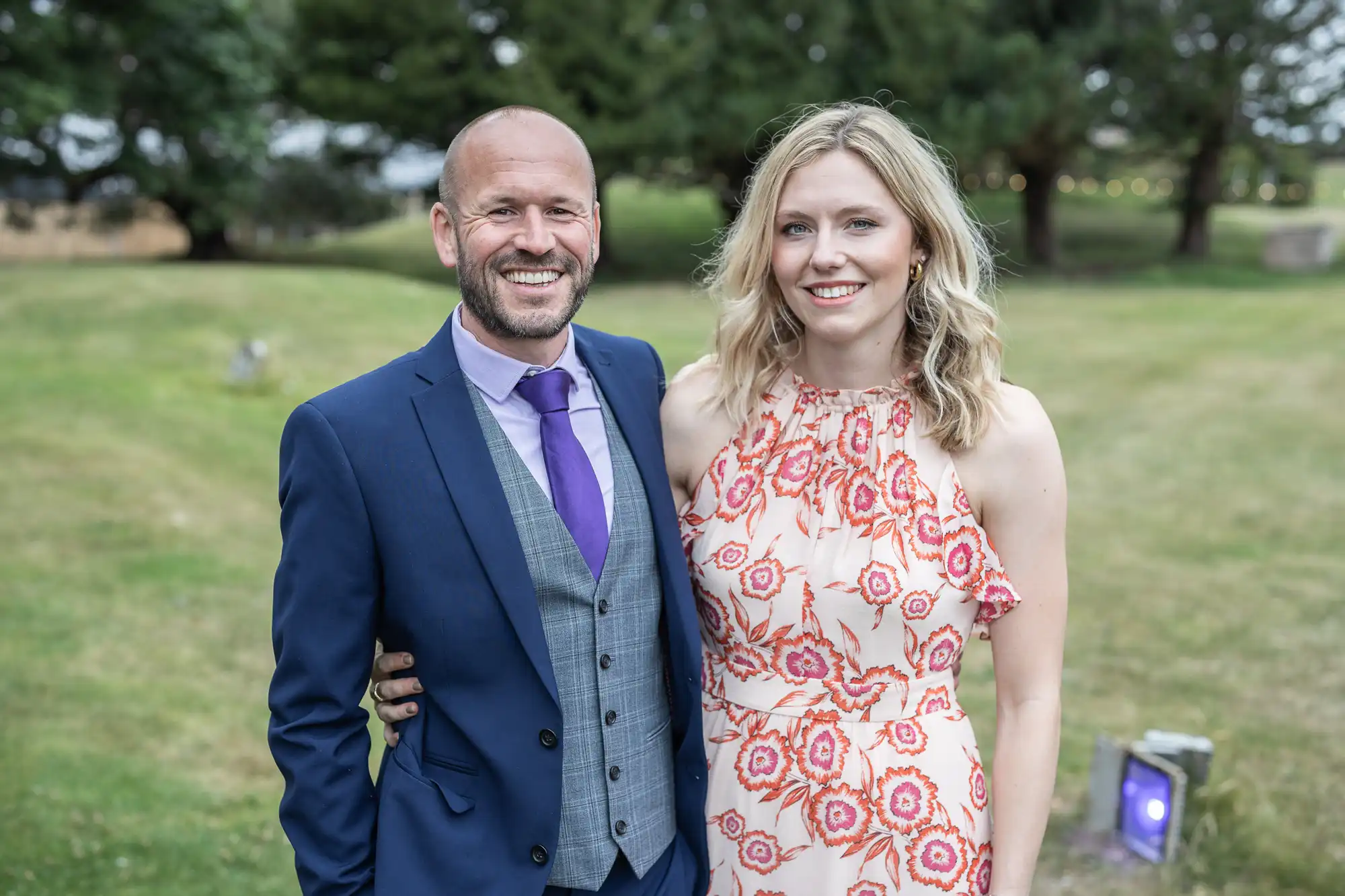 A man in a blue suit and a woman in a floral dress stand together outdoors on a grassy area, smiling at the camera. Trees can be seen in the background.