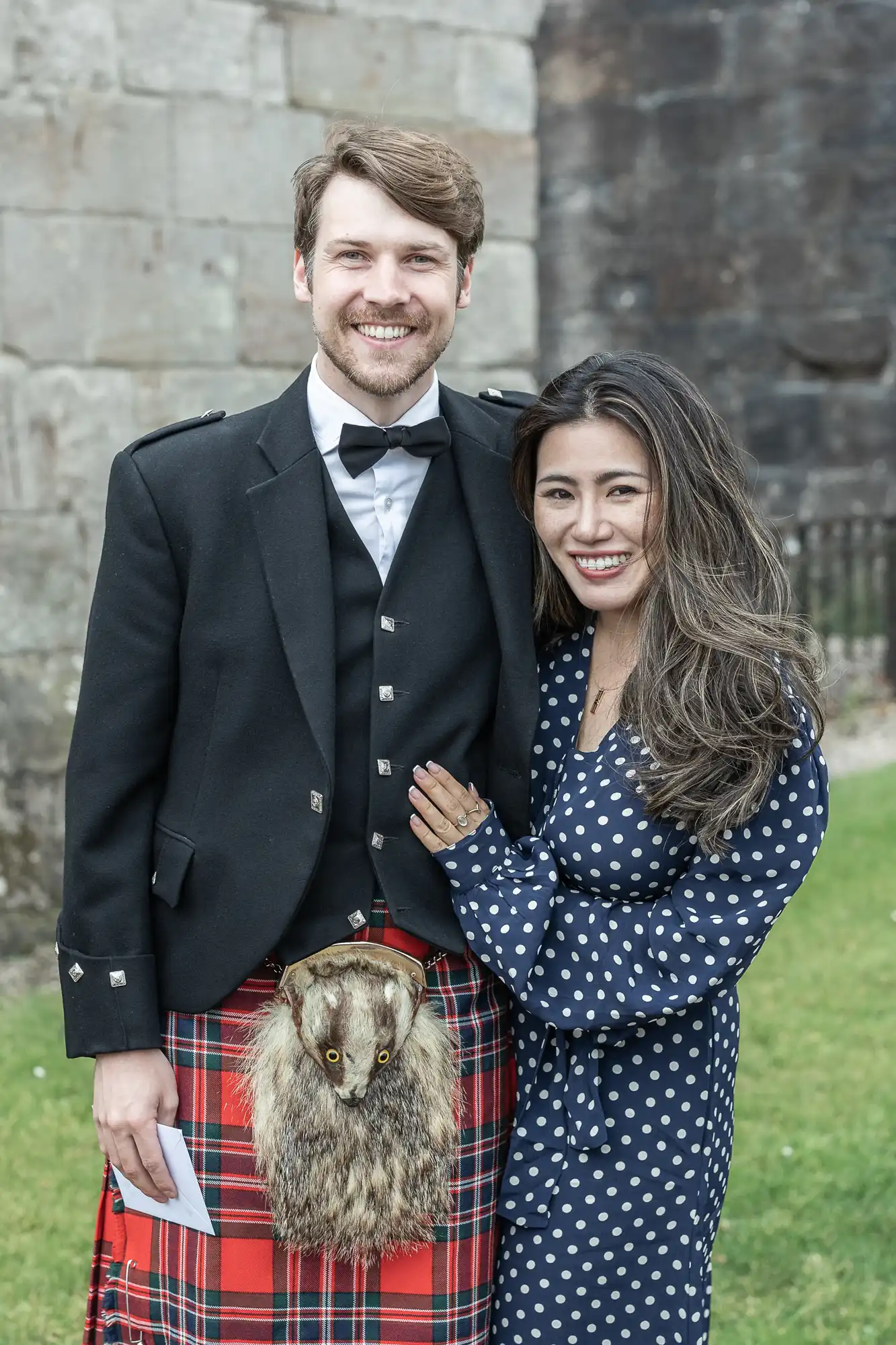 A man and woman stand together outside, smiling. The man is wearing Scottish traditional attire, including a kilt and sporran. The woman is wearing a blue polka-dot dress.