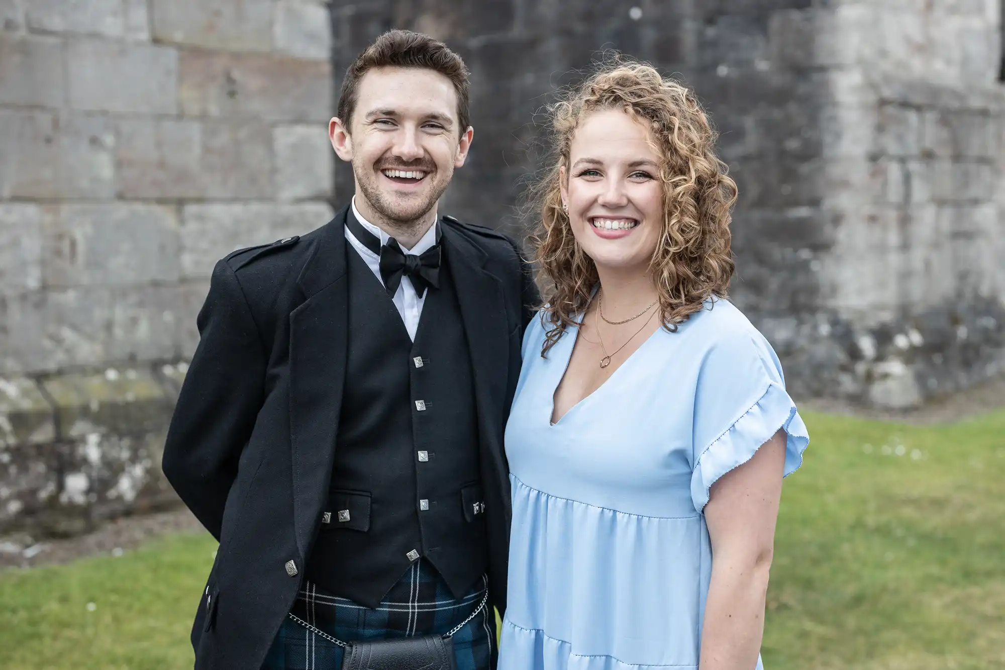 A man in a black traditional Scottish outfit and a woman in a light blue dress stand together, smiling, in front of an old stone building.