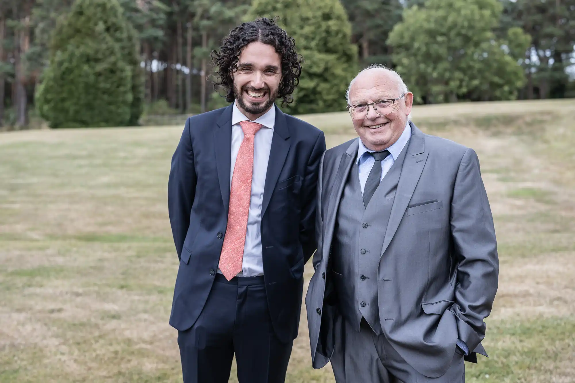 Two men, one younger and one older, dressed in suits, stand side by side smiling in an outdoor setting with trees and grass in the background.