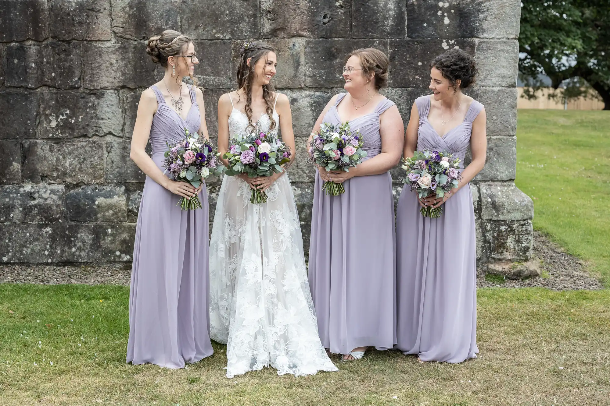 A bride in a white gown and three bridesmaids in lavender dresses stand together outside, holding flower bouquets and smiling at each other. They are positioned near a stone wall and a grassy area.