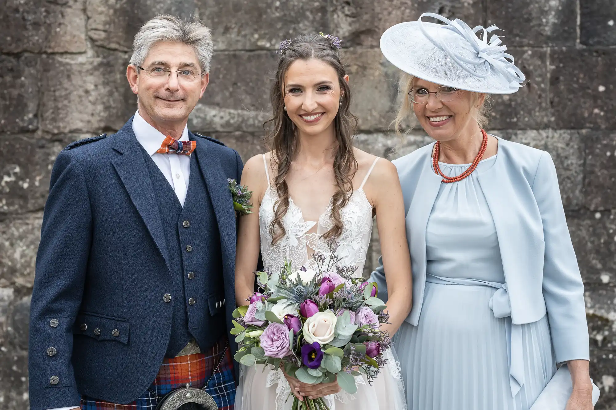 A bride in a white dress holding a bouquet poses between an older man in a kilt and an older woman wearing a blue dress and hat against a stone background.