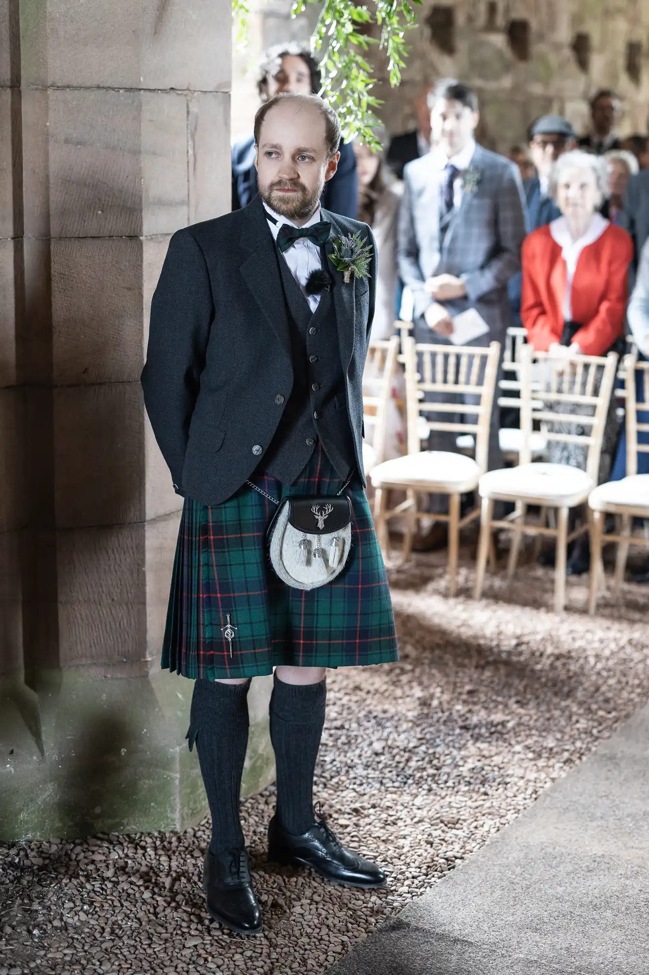 A man in a dark jacket and tartan kilt stands by a stone wall in a room with guests seated in the background.