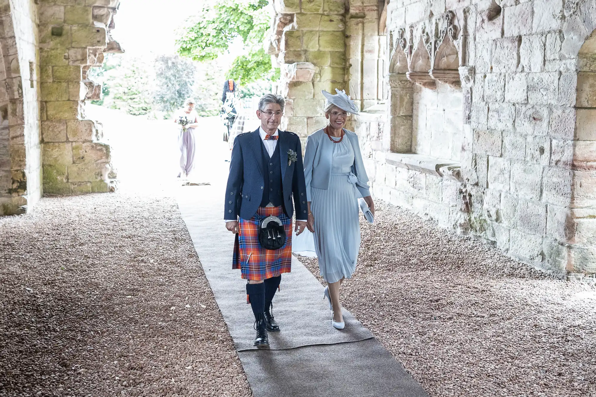 Two people, dressed in formal attire, walk down a pathway beside a stone building. The man wears a kilt, and the woman is in a light blue dress and hat.