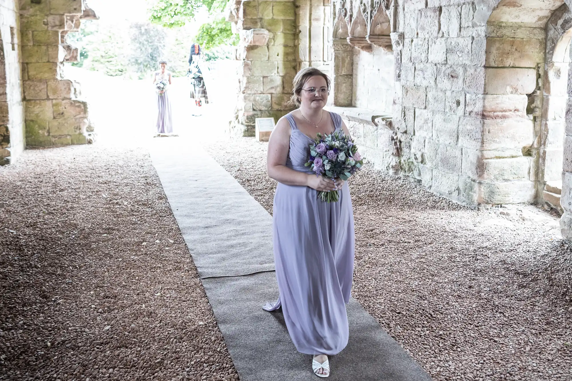 A woman in a lavender dress holding a bouquet walks down an aisle in a stone building. Another person in a similar dress and a figure in black are in the background.