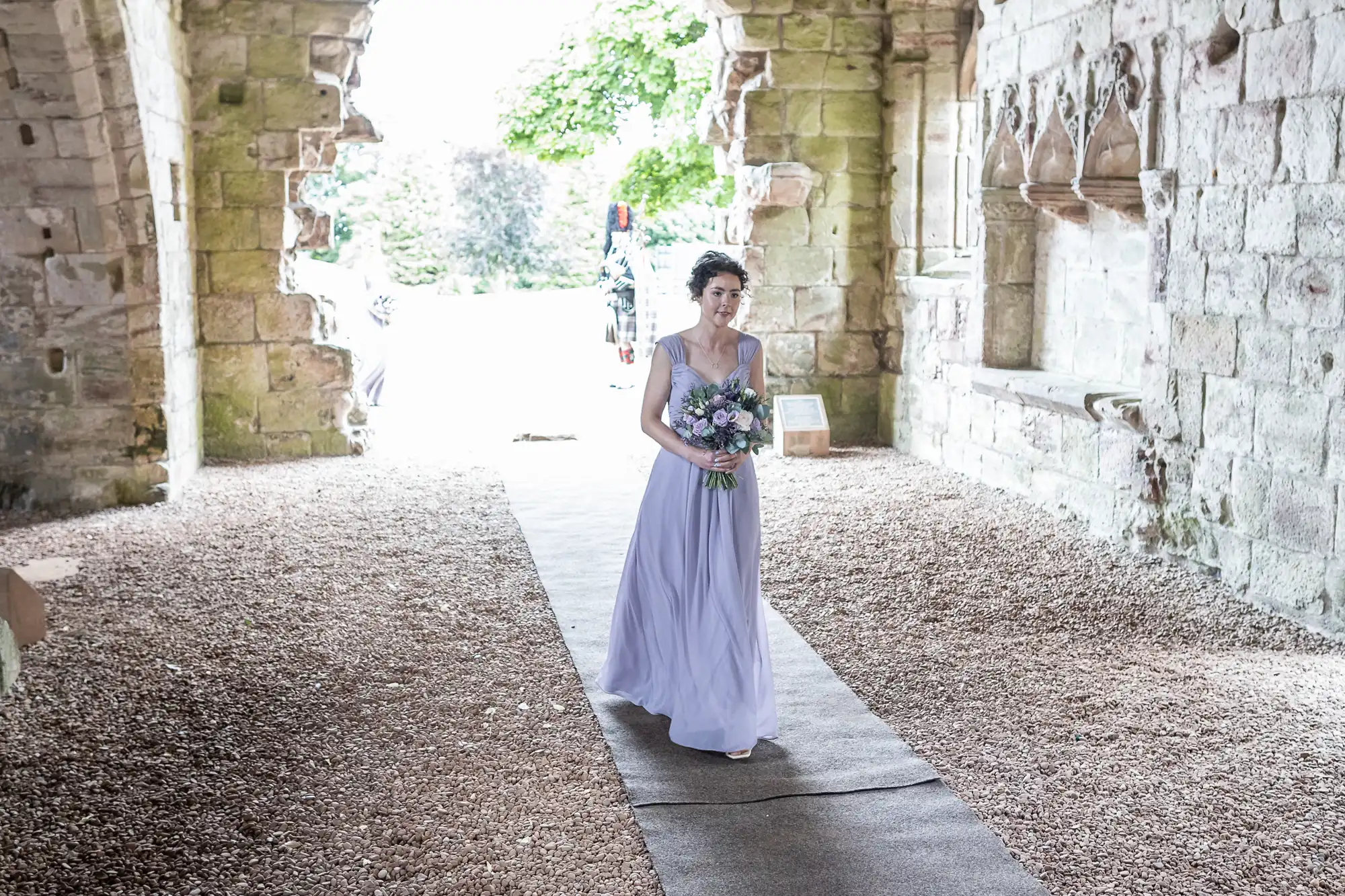 A woman in a light purple dress walks down a gravel path in a stone building, holding a bouquet of flowers.