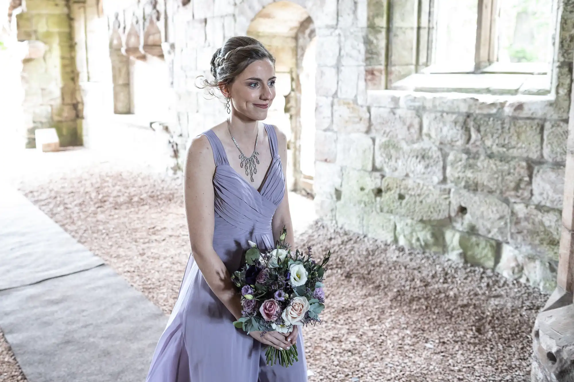 A woman in a light purple dress holds a bouquet of flowers while standing in a stone building with arched windows.