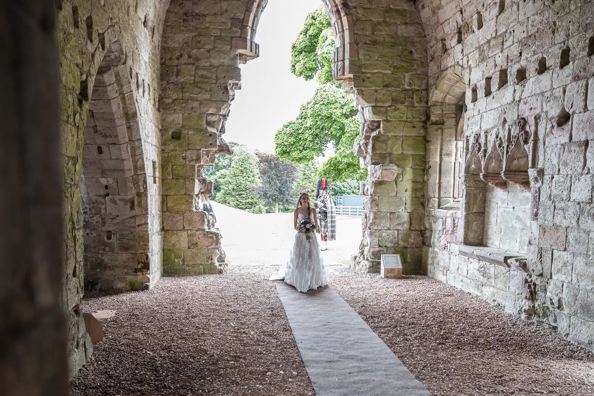 A bride in a white gown stands under an ancient stone archway, holding a bouquet, with greenery and trees visible through the opening behind her.