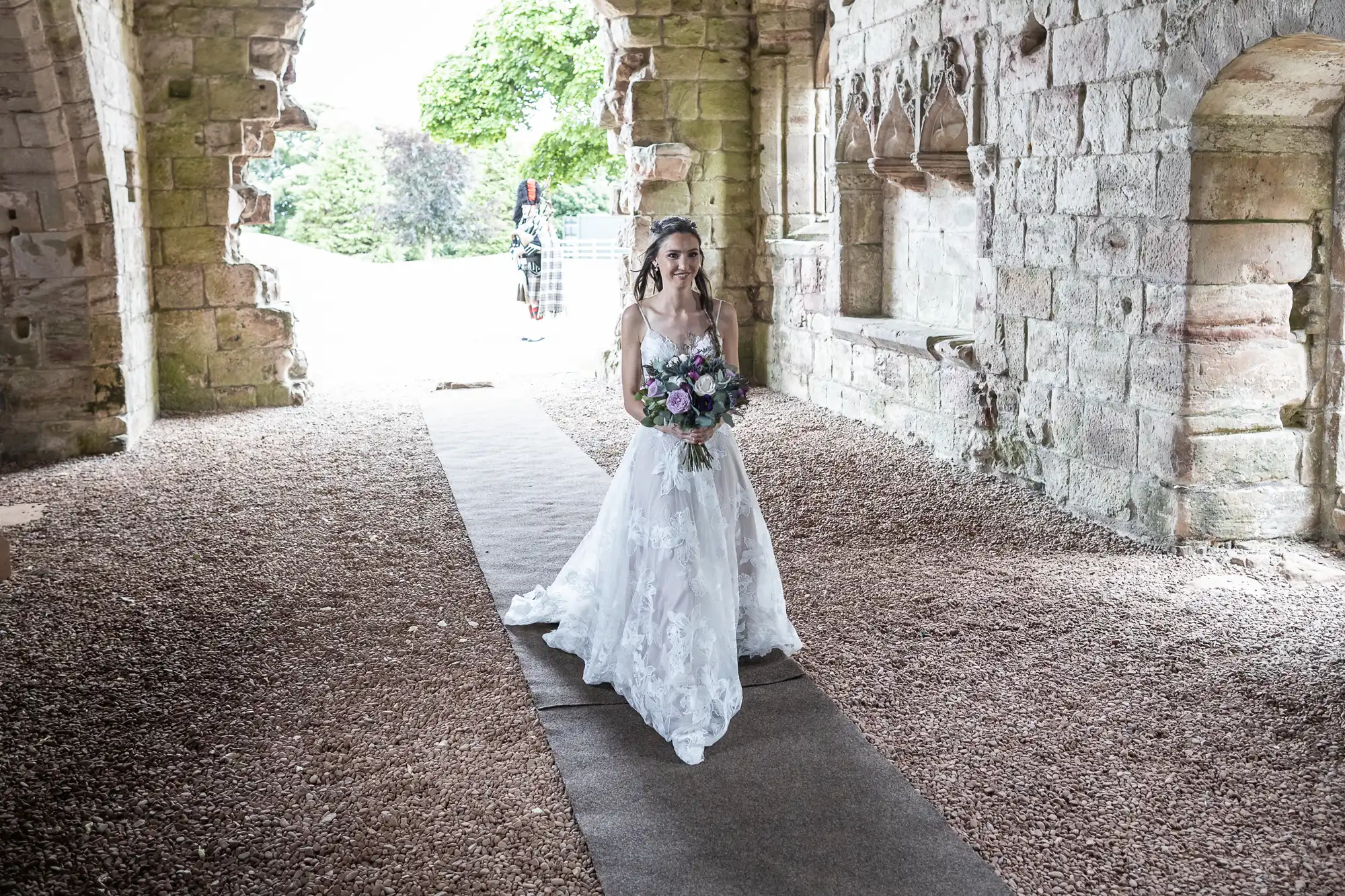 A bride in a white dress holds a bouquet of flowers, walking down an aisle in an old stone building.