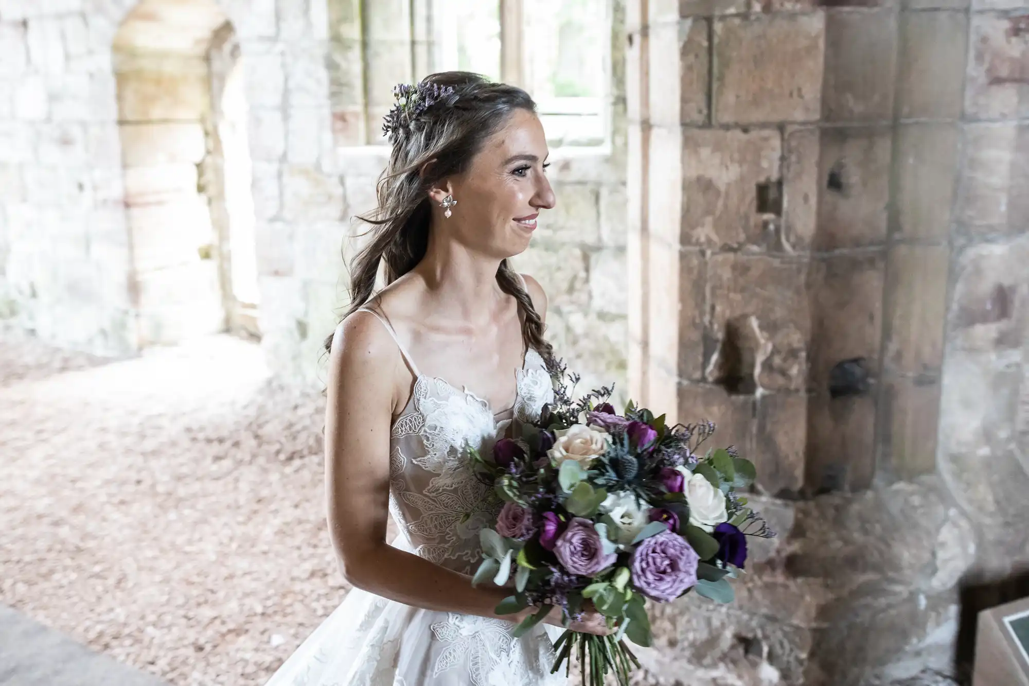 A bride in a lace wedding dress holds a bouquet of flowers while standing in an old stone building.