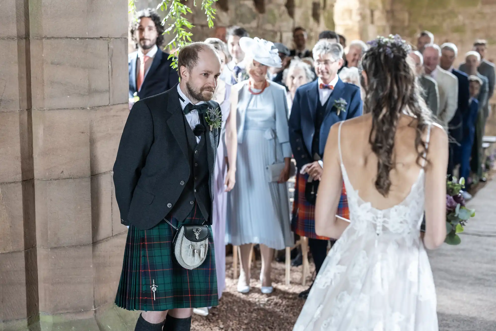 A groom in a kilt stands facing the bride during a wedding ceremony, with guests in the background.