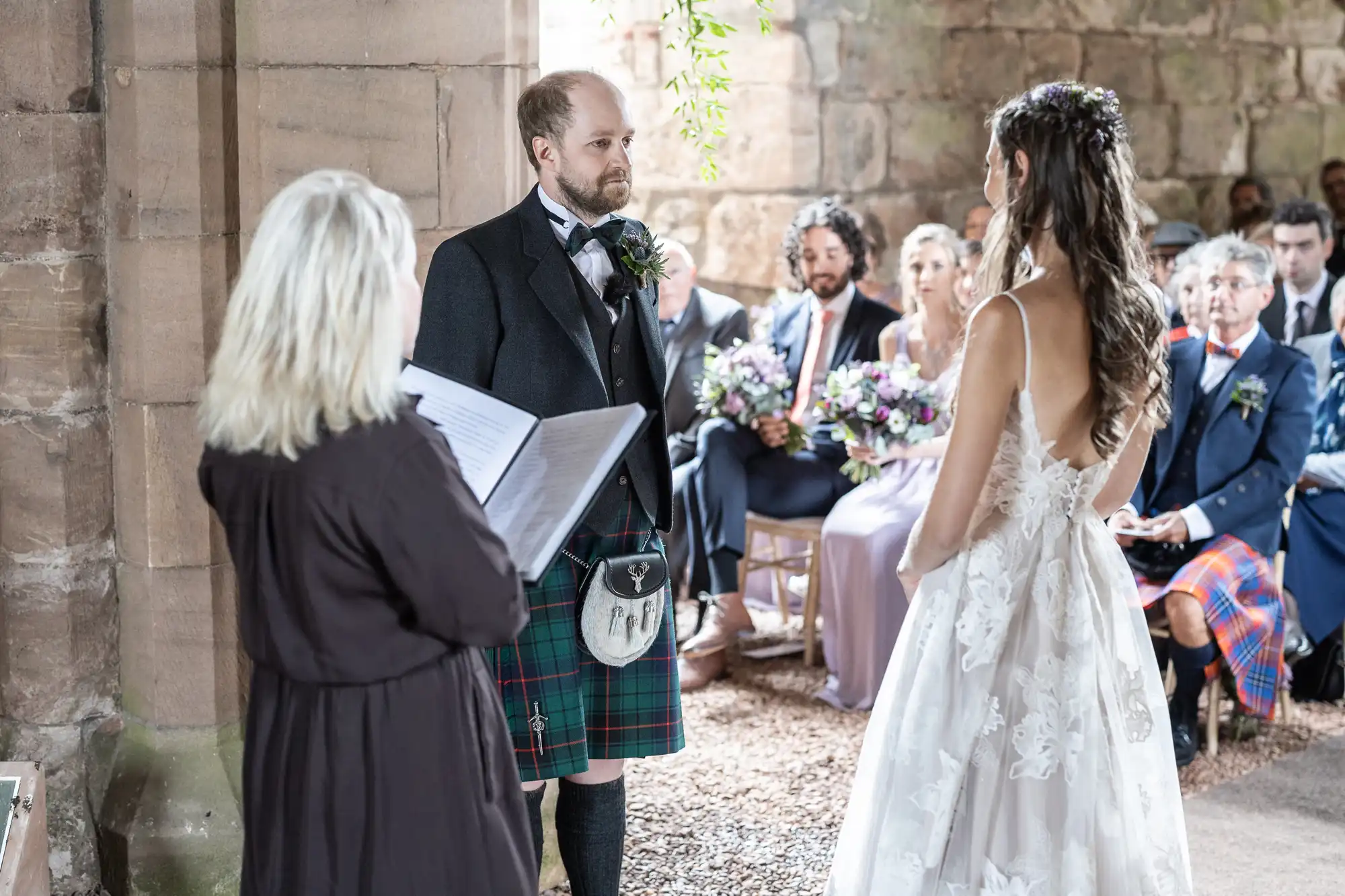 A bride and groom, with the groom in a kilt, stand facing each other during a wedding ceremony while an officiant reads from a book. Wedding guests are seated in the background.