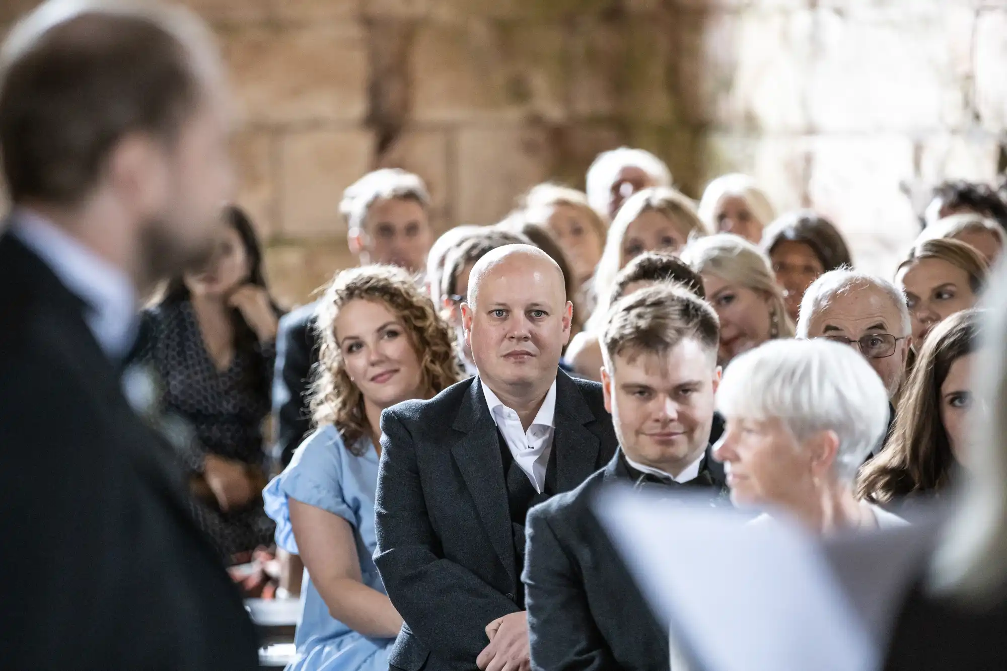 A group of people, dressed in formal attire, attentively watch a person speaking at an indoor event.