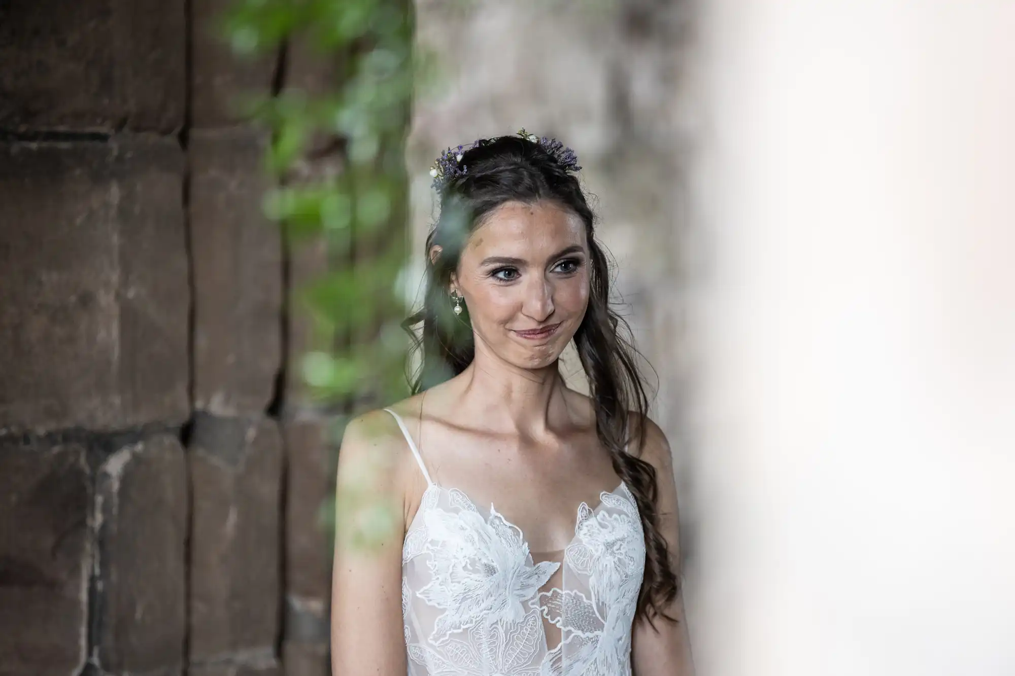 A woman in a white lace dress stands smiling, wearing a floral hairpiece, with a stone wall in the background.