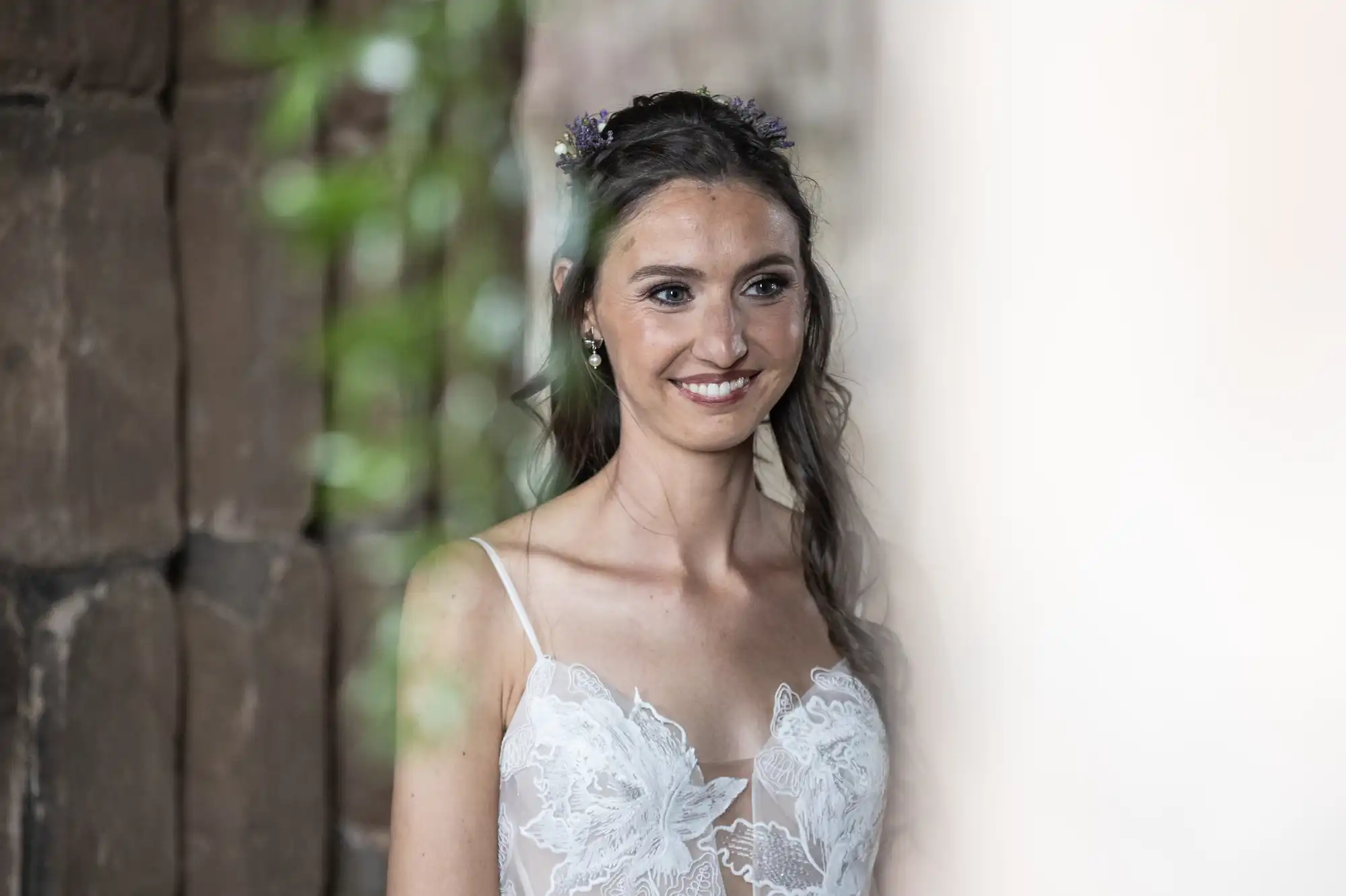 A woman in a white lace bridal gown with floral headpiece smiles in front of a stone wall.