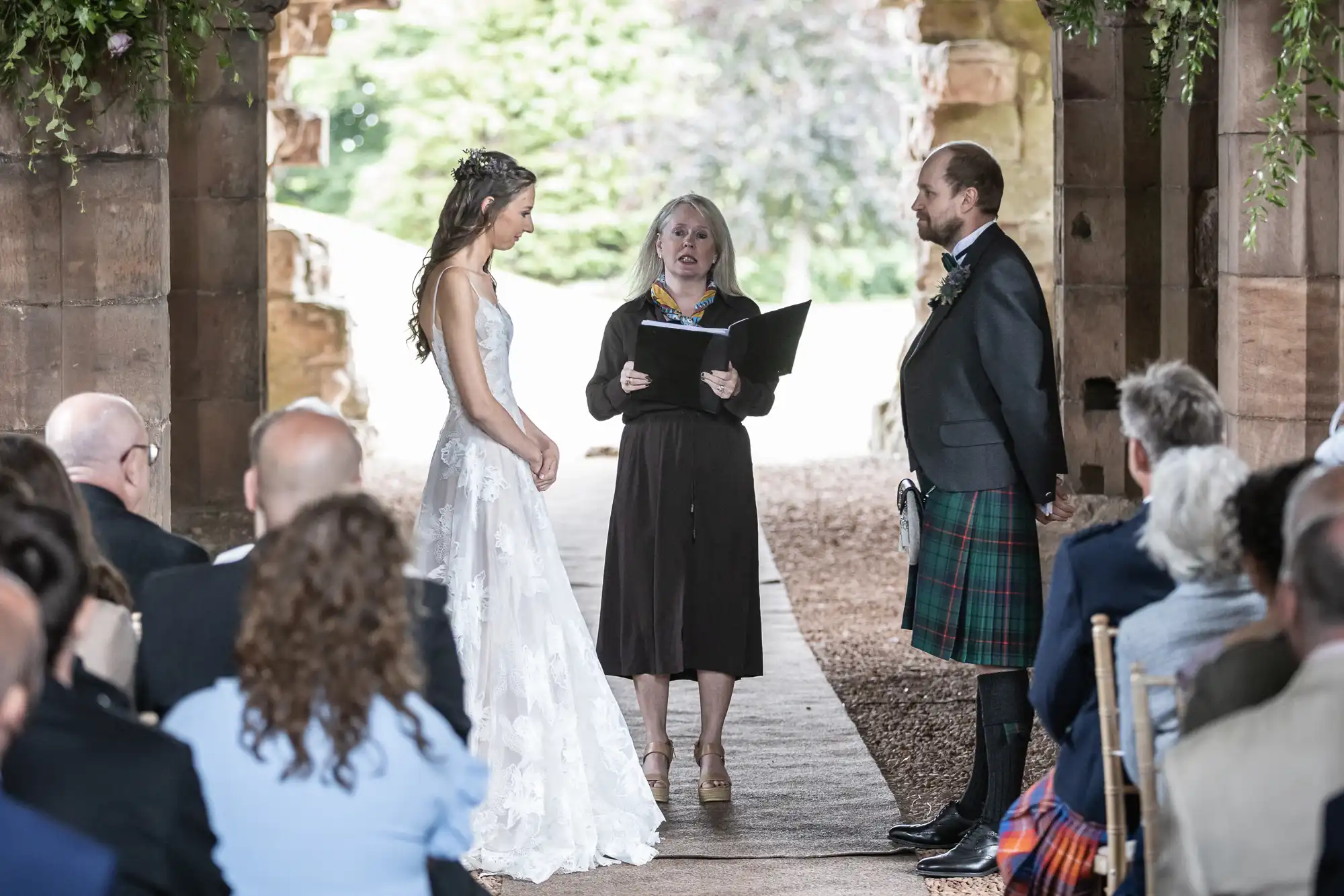 A bride and groom stand facing each other during an outdoor wedding ceremony officiated by a woman, with guests seated and watching.