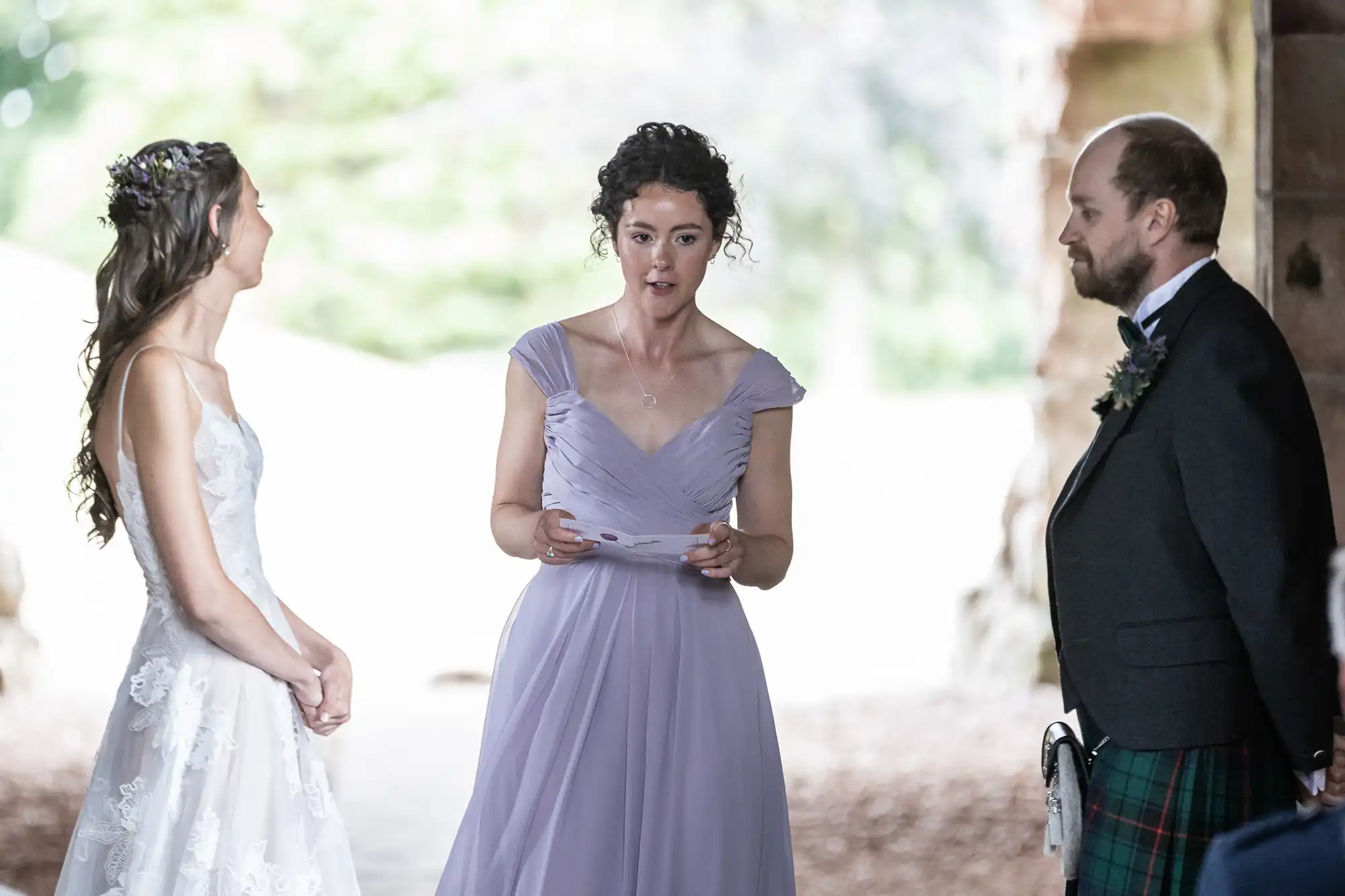 Three people stand together. One woman in a white dress listens, another woman in a light purple dress speaks, holding a paper, and a man in a suit with a kilt looks on. They are outdoors.