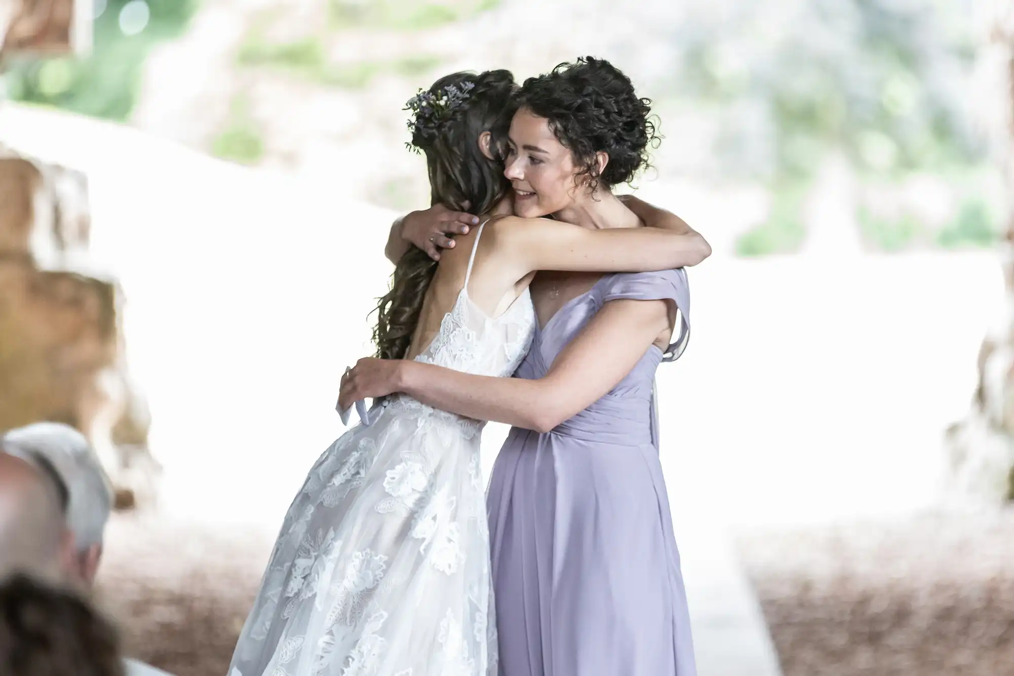 Two women embrace affectionately, one in a white lace wedding dress and the other in a lavender gown, during a outdoor ceremony.