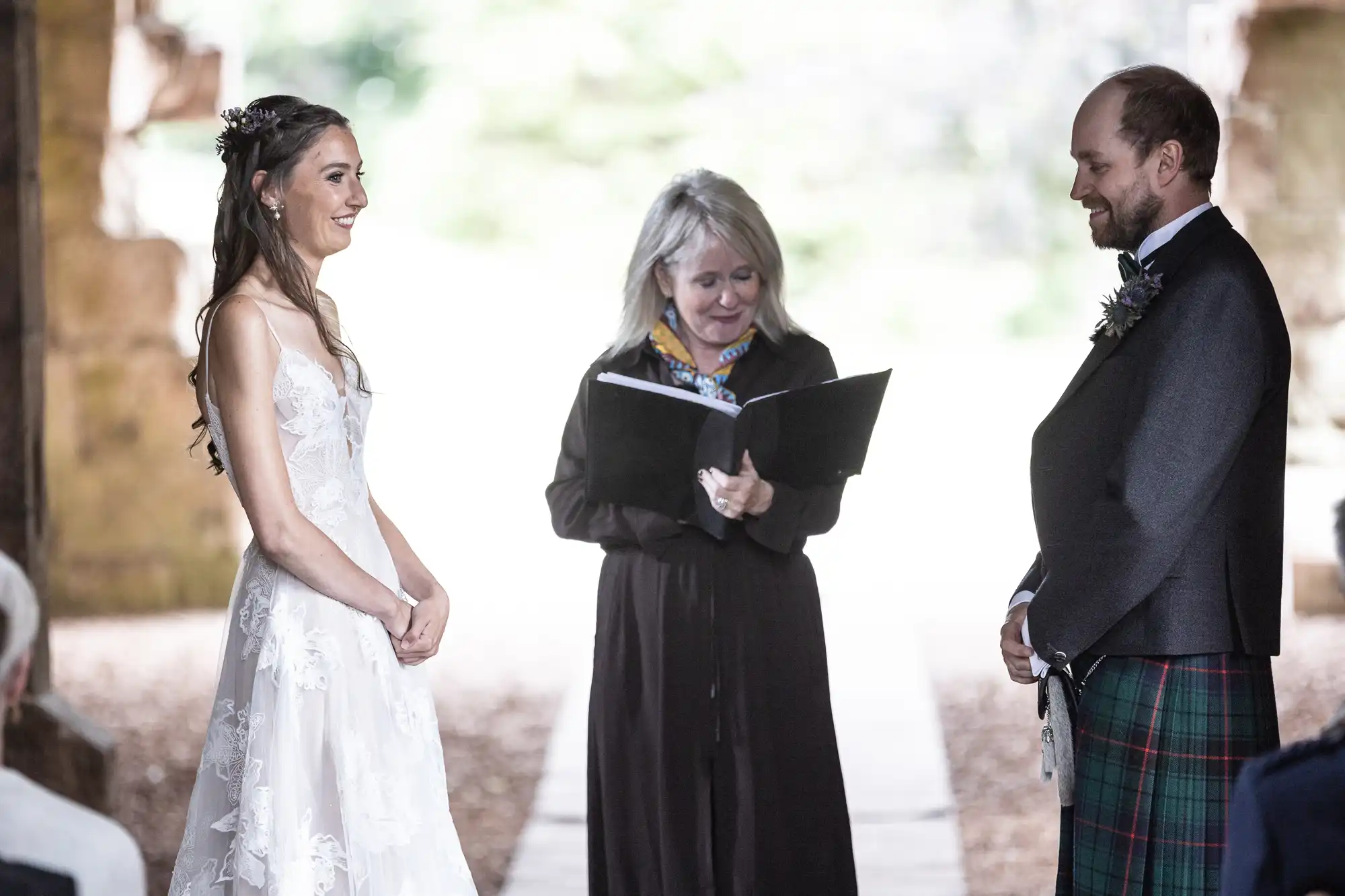 A couple stands facing each other during their wedding ceremony while an officiant reads from a book. The bride wears a white dress, and the groom is dressed in a kilt and formal attire.