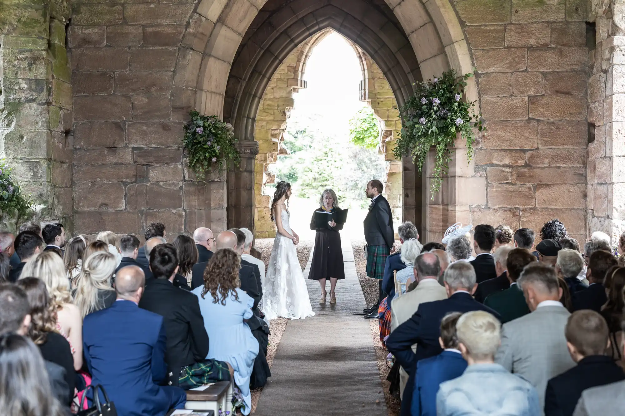 A bride and groom stand before an officiant in an arched stone structure, surrounded by seated guests, during a wedding ceremony.