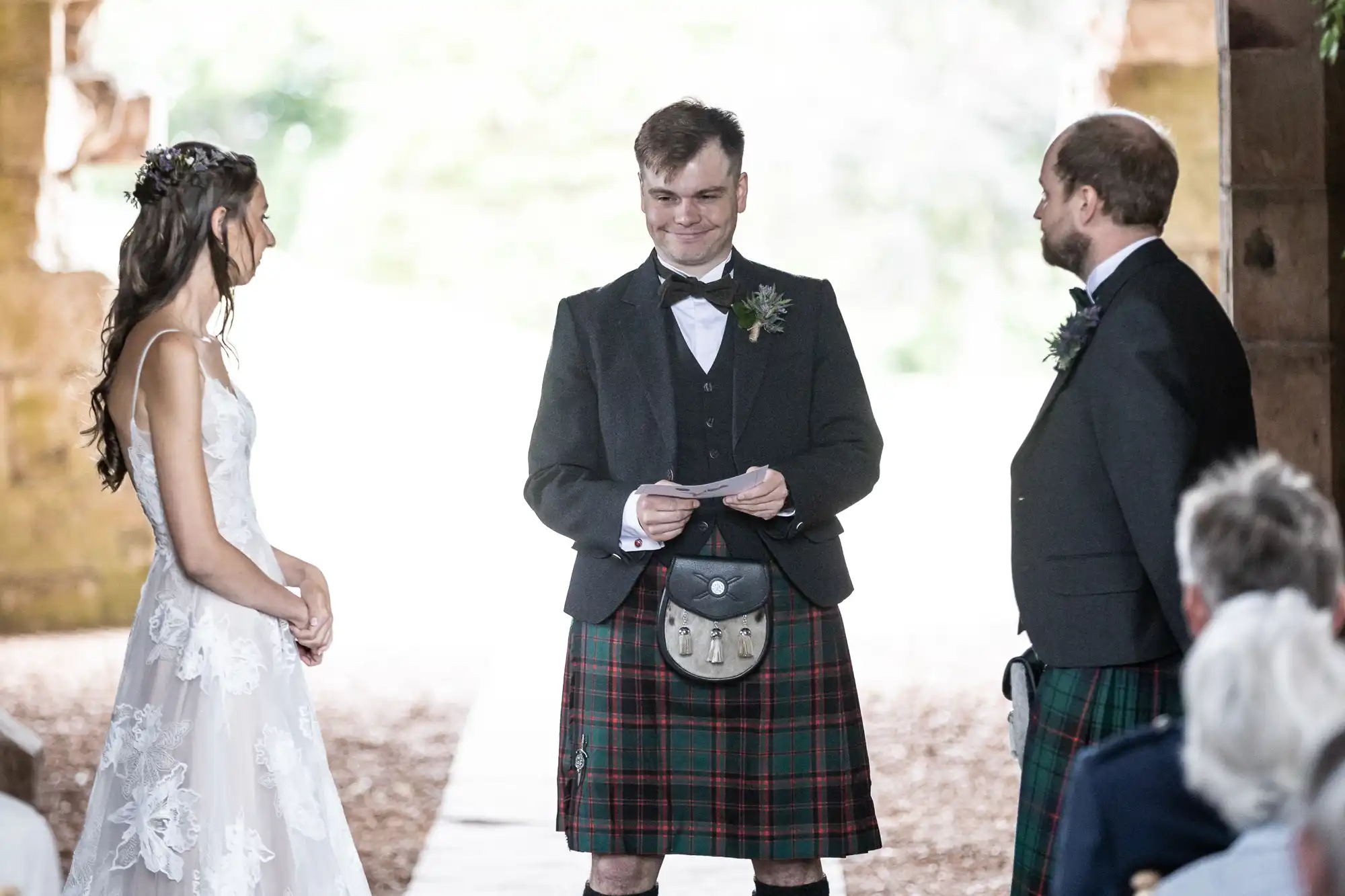 Three people dressed in formal attire, two men in kilts and a woman in a white dress, standing and smiling during a ceremony.