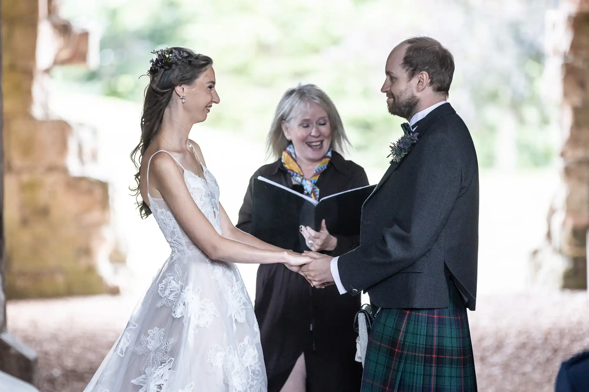 A bride and groom hold hands and smile at each other during their wedding ceremony, with an officiant standing behind them, holding a book and appearing to speak.