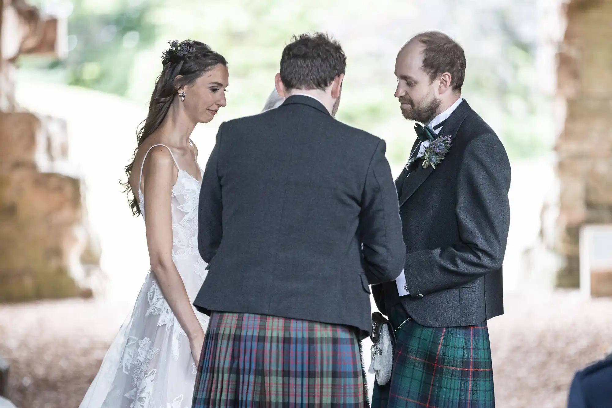 A bride and groom stand facing an officiant during their wedding ceremony, with the groom and the officiant both wearing traditional Scottish kilts.