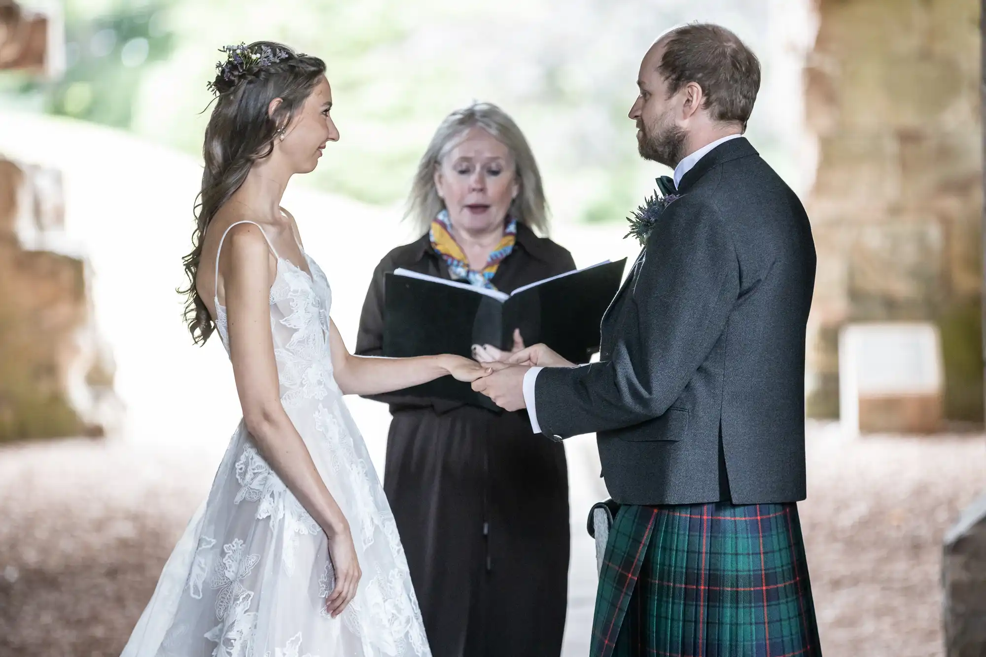 A bride and groom exchange vows while holding hands, with an officiant standing behind them reading from a book, all in a well-lit, outdoor setting.