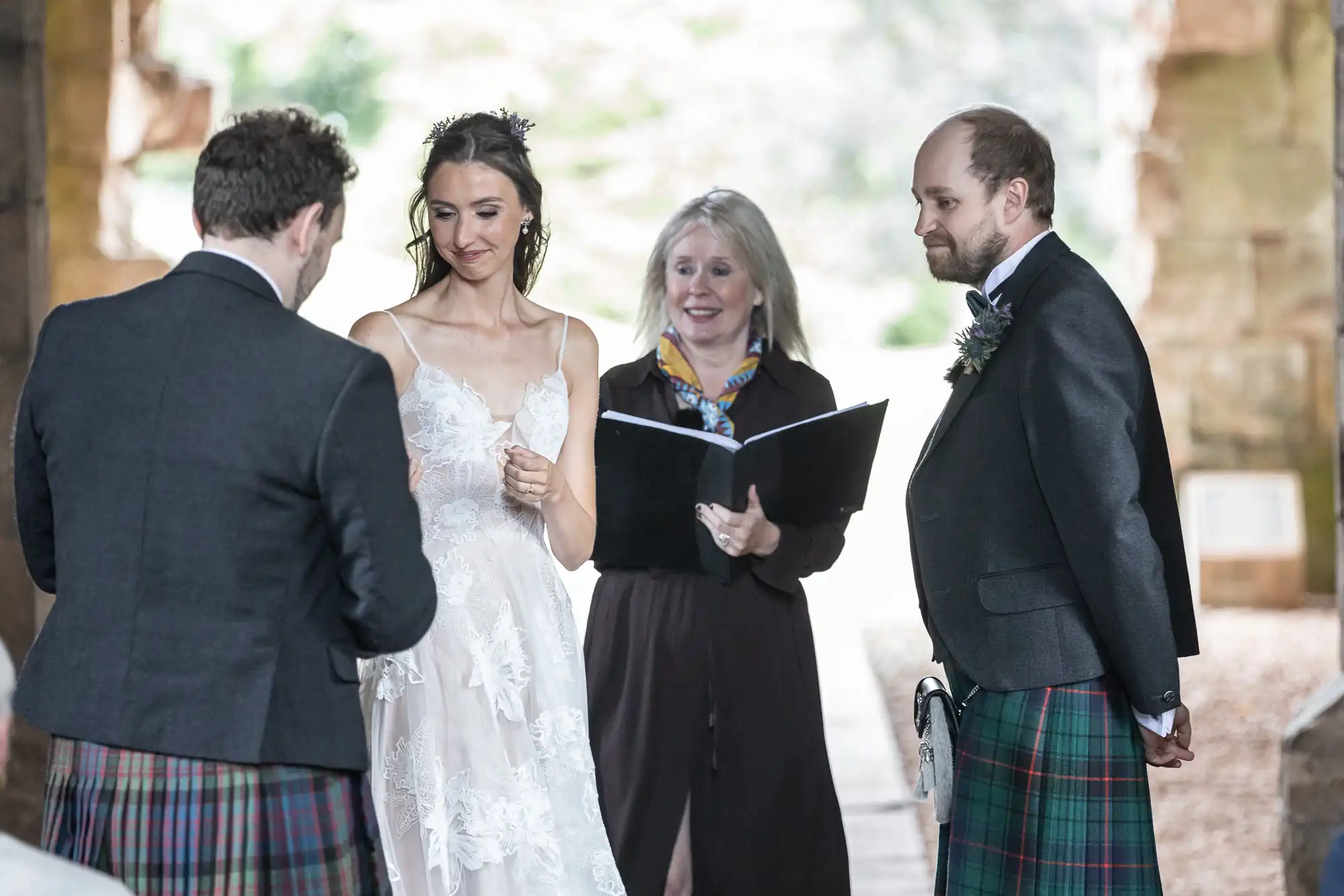 A bride and a groom in kilts stand facing another man in a kilt. A woman holding an open book stands between them. The setting appears to be an outdoor location with stone structures in the background.