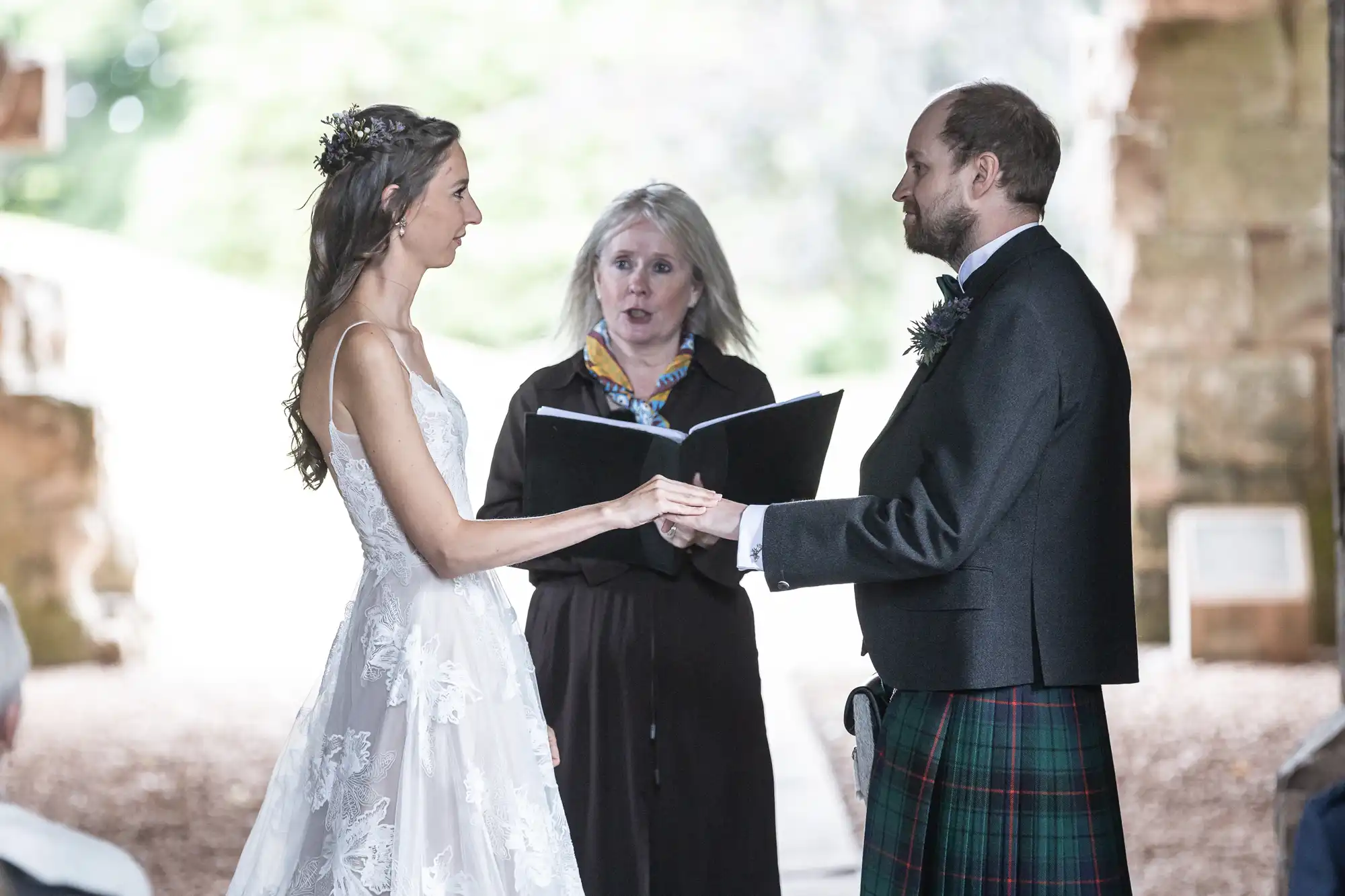 A bride and groom holding hands, facing each other, while an officiant stands in between them, reading from a book during their outdoor wedding ceremony.