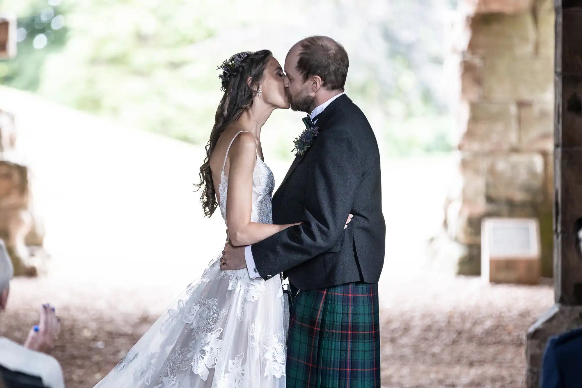 A couple shares a kiss during their wedding ceremony. The bride wears a white lace dress and the groom is dressed in a suit with a tartan kilt. They stand in a rustic, open stone structure.