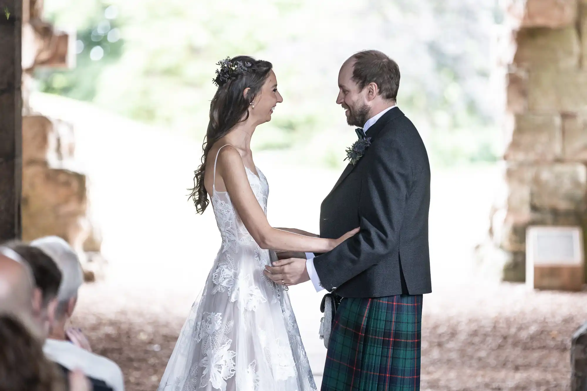 A bride in a white lace gown and a groom in a dark jacket with a tartan kilt stand facing each other, smiling, hands joined, in an outdoor setting during a wedding ceremony.