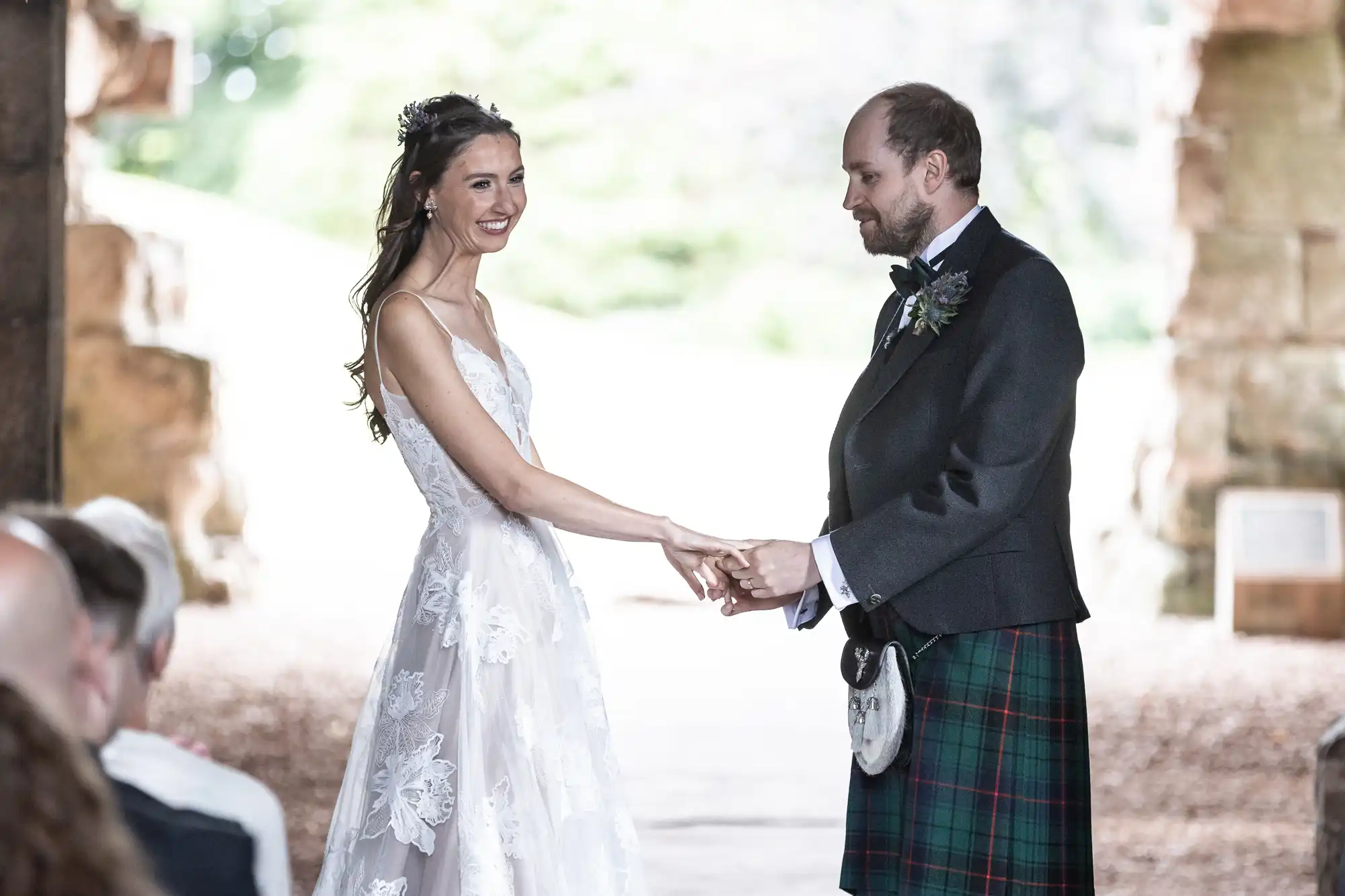 A bride in a white lace gown and a groom in a tartan kilt hold hands during their wedding ceremony, standing amid historic stone ruins in a wooded area.