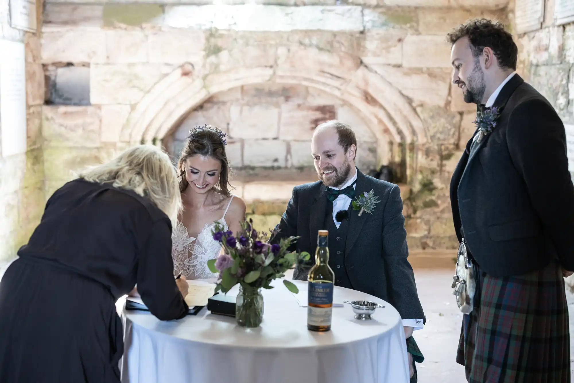 A bride and groom sit at a table while a woman signs a document. A man in a kilt stands beside them. A bottle of whiskey and a floral arrangement are on the table.