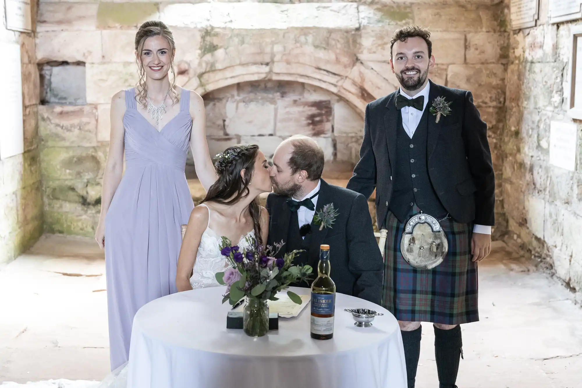 A bride and groom share a kiss while seated at a table with a bouquet and bottle. Two people, one in a dress and the other in a kilt, stand on either side, smiling at the camera.