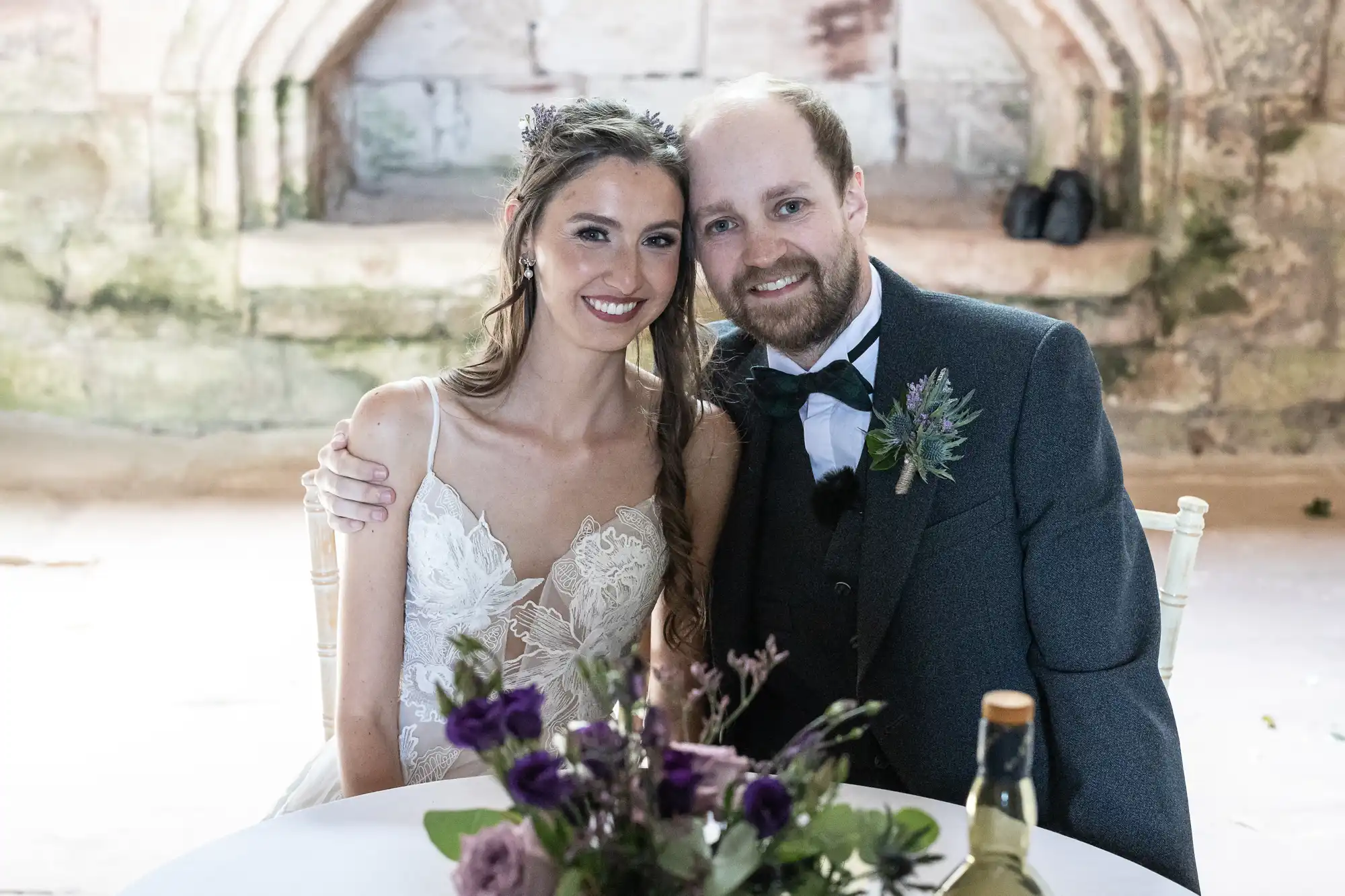 A smiling couple in wedding attire poses together at a table adorned with flowers and a wine bottle, set against a rustic stone backdrop.