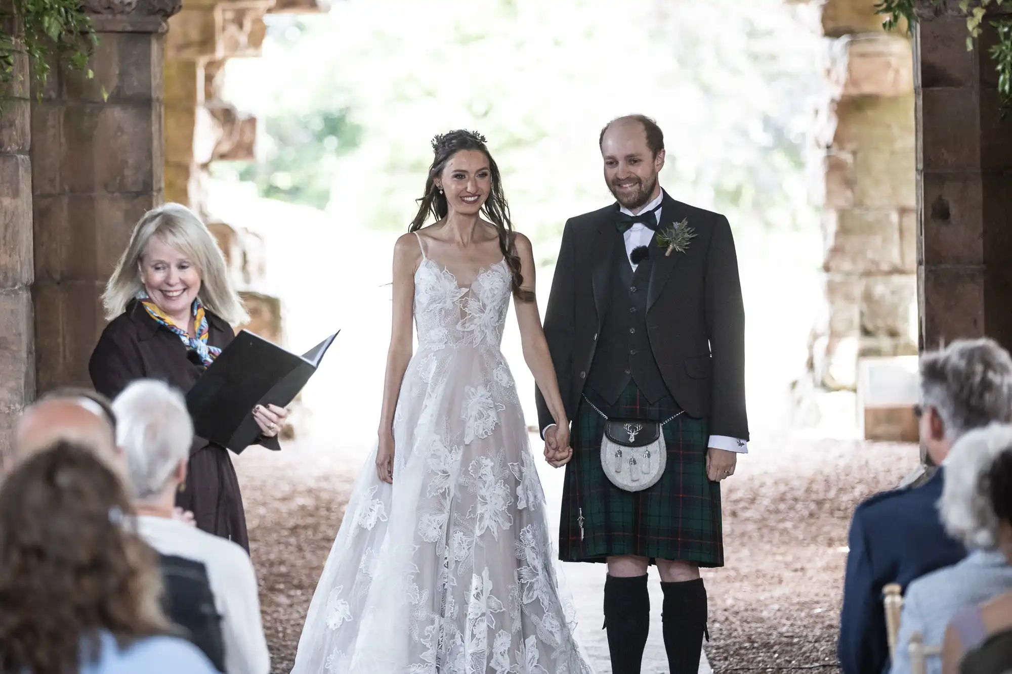 A bride in a white gown and groom in a kilt stand hand in hand, smiling during their outdoor wedding ceremony with an officiant and guests.