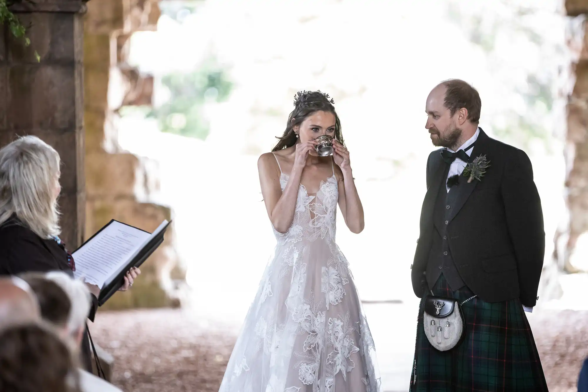 A bride and groom in wedding attire participate in a ceremonial moment with an officiant standing nearby, holding an open book.