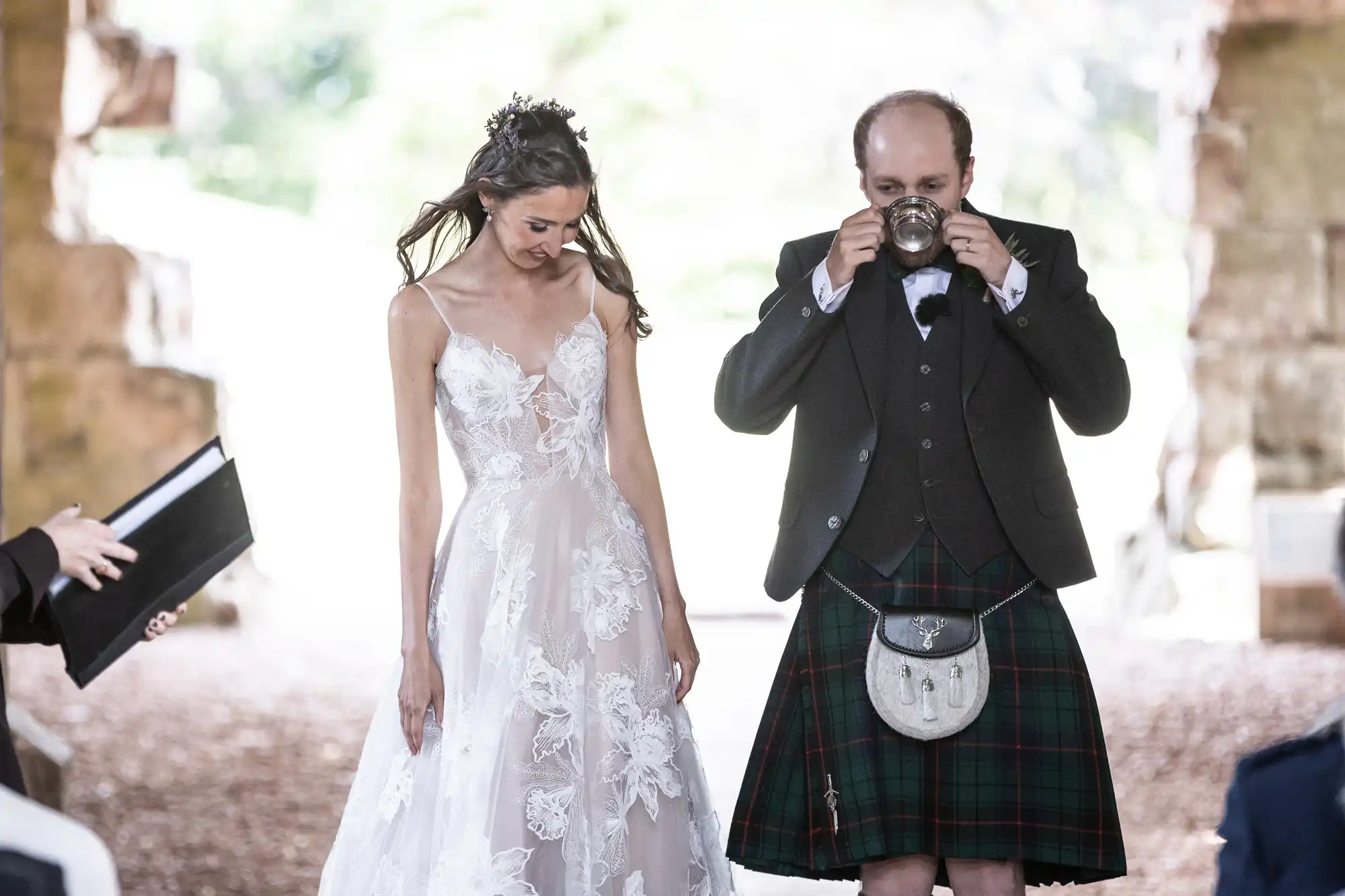 Bride in white floral wedding dress and groom in a kilt stand together. The groom drinks from a cup while the bride looks down smiling. Stone wall visible in the background.