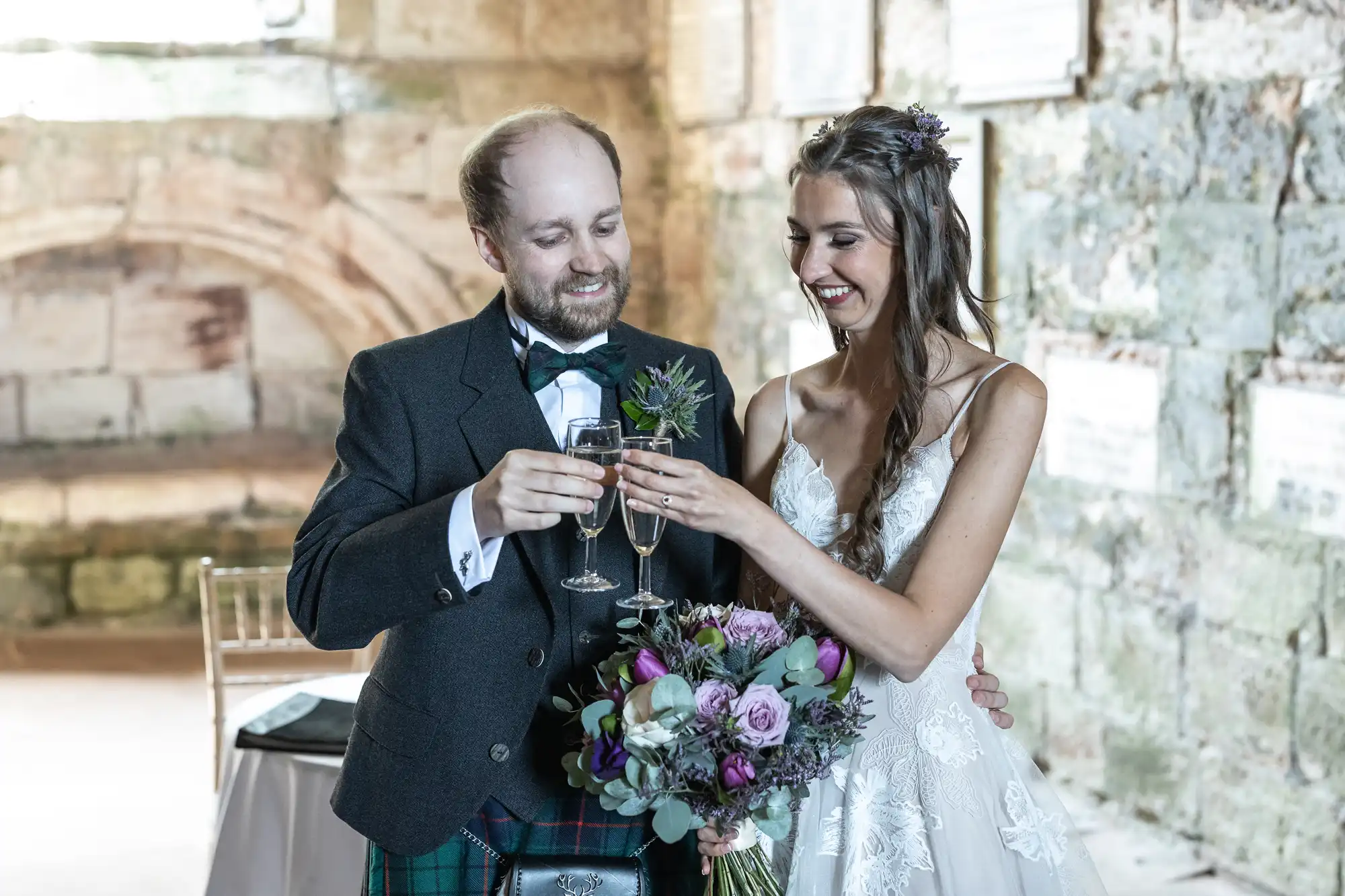 A couple dressed in wedding attire clink glasses in a toast. The bride holds a bouquet of flowers, and both are smiling. They stand in a rustic, stone-walled setting.