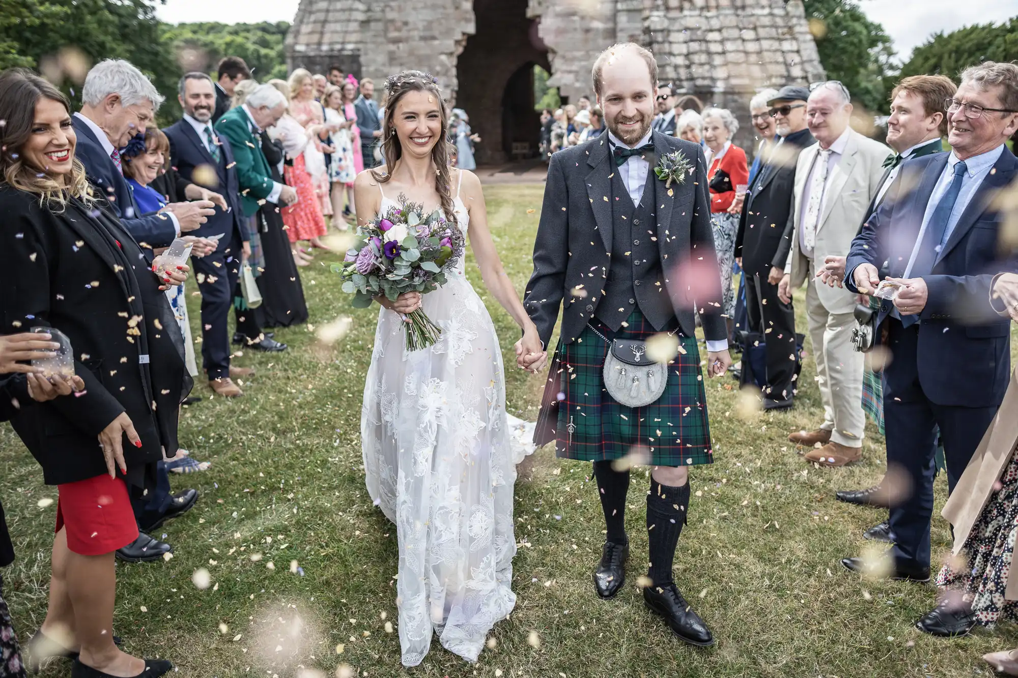 A bride and groom walk hand-in-hand at an outdoor wedding ceremony, smiling as guests throw confetti. The groom is wearing a kilt, and the bride is holding a bouquet of flowers.