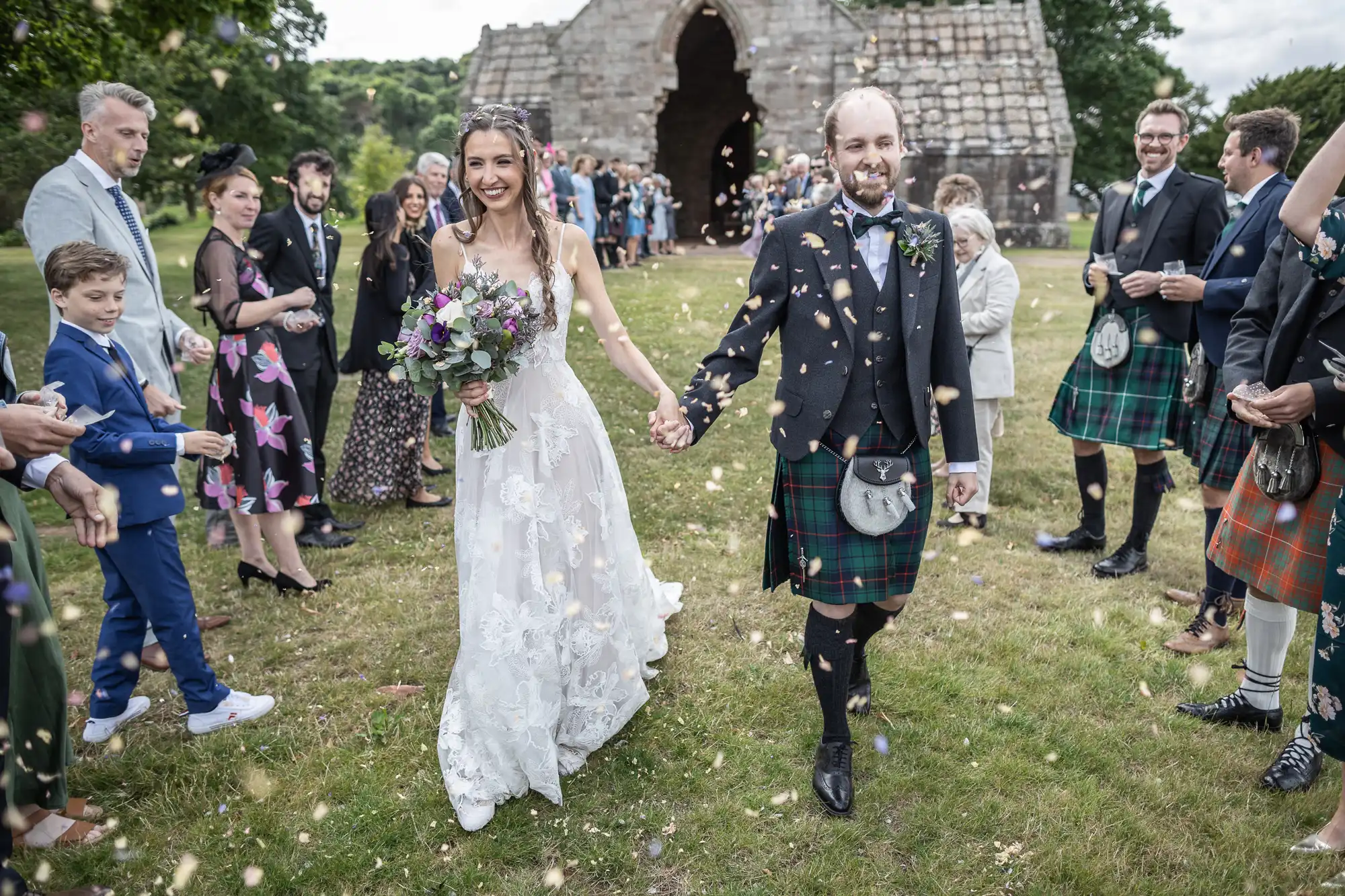 Bride and groom walk hand in hand under a shower of confetti. Guests dressed in various attires, including kilts, look on. A stone building is visible in the background.