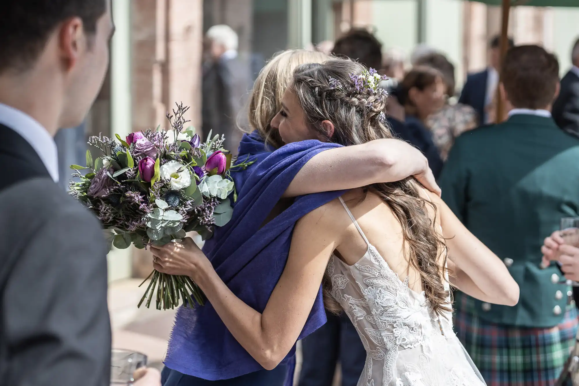 A bride in a wedding dress hugs another person holding a bouquet of purple and white flowers during an outdoor event.