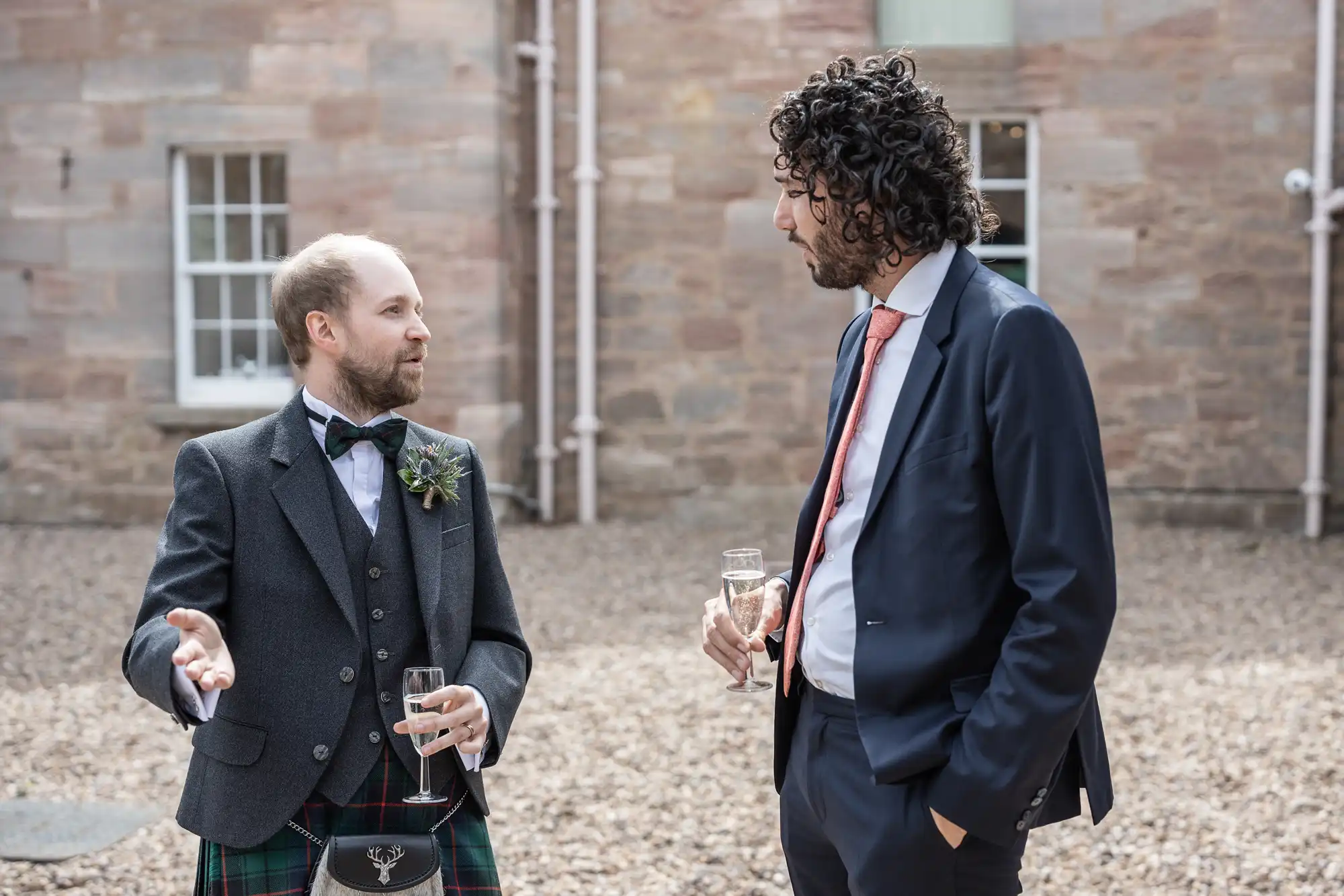 Two men engaged in conversation, one wearing a traditional Scottish kilt and jacket, the other in a blue suit and red tie. Both are holding glasses, standing outside a stone building with gravel ground.