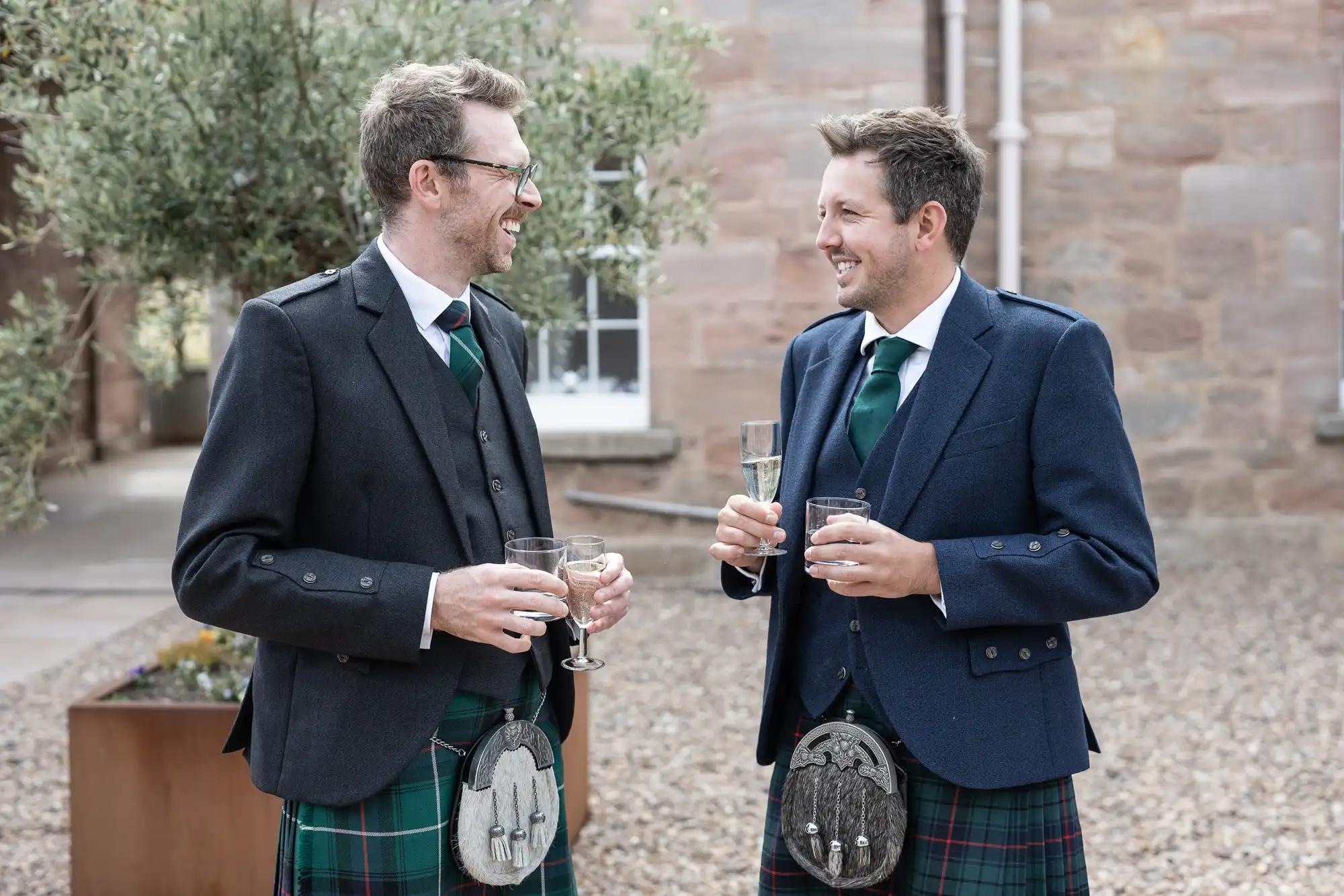 Two men in formal Scottish kilts and jackets smile and hold drinks while standing outside near a stone building and an olive tree.