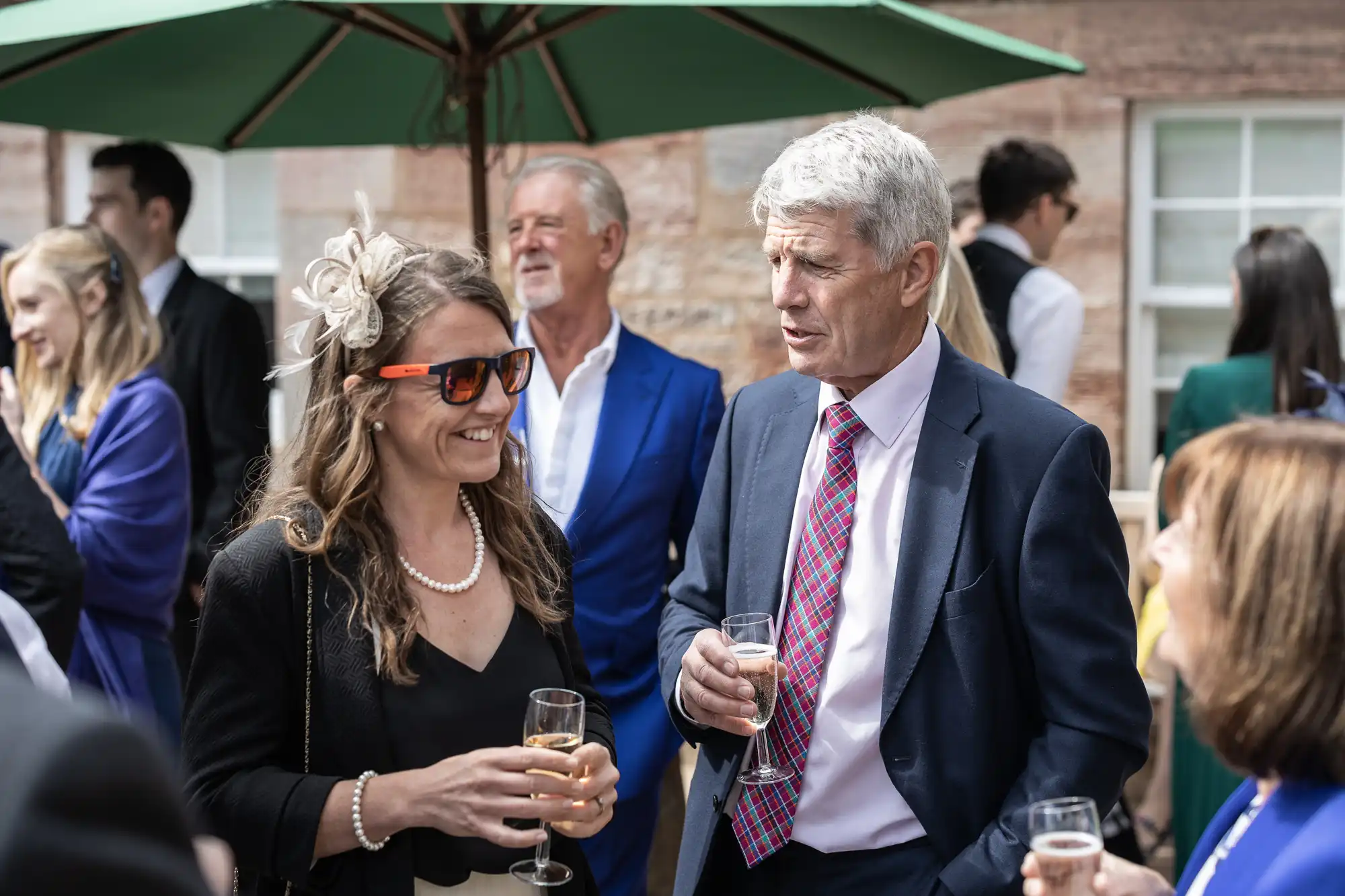 A group of people in formal attire are socializing outdoors under a green umbrella, holding glasses of champagne.