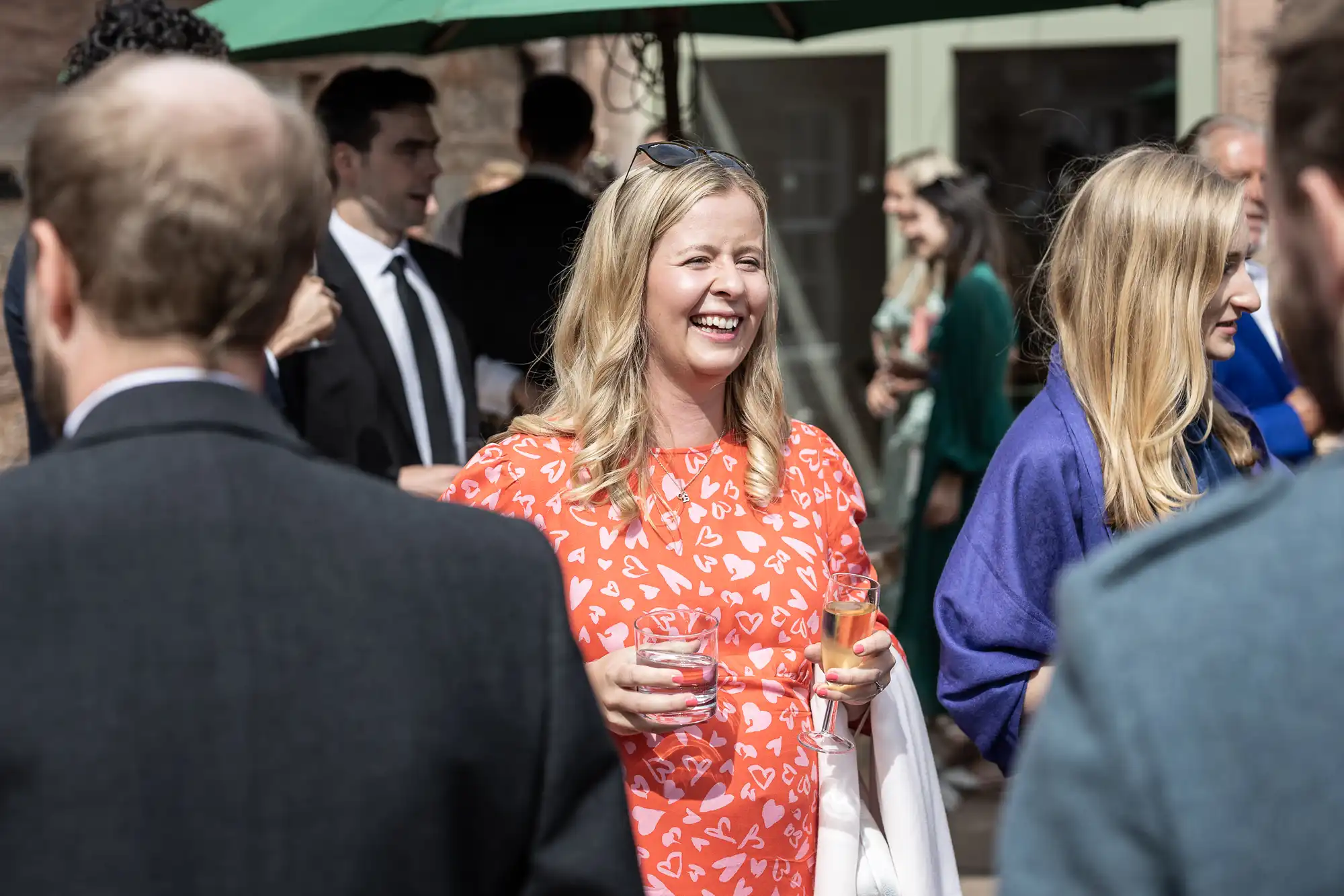 A woman in an orange dress with white patterns holding a drink smiles at an outdoor social gathering. Several other people, both men and women, are interacting in the background.