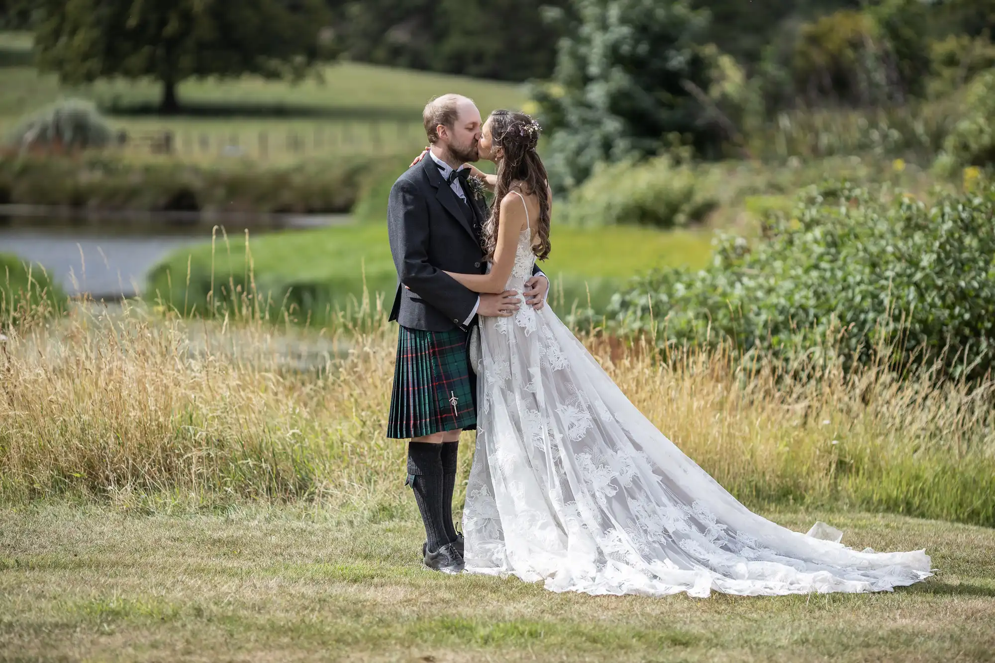 A couple dressed in wedding attire share a kiss outdoors. The man is in a traditional kilt, and the woman is in a long white dress with a flowing train. They are standing in a grassy field.