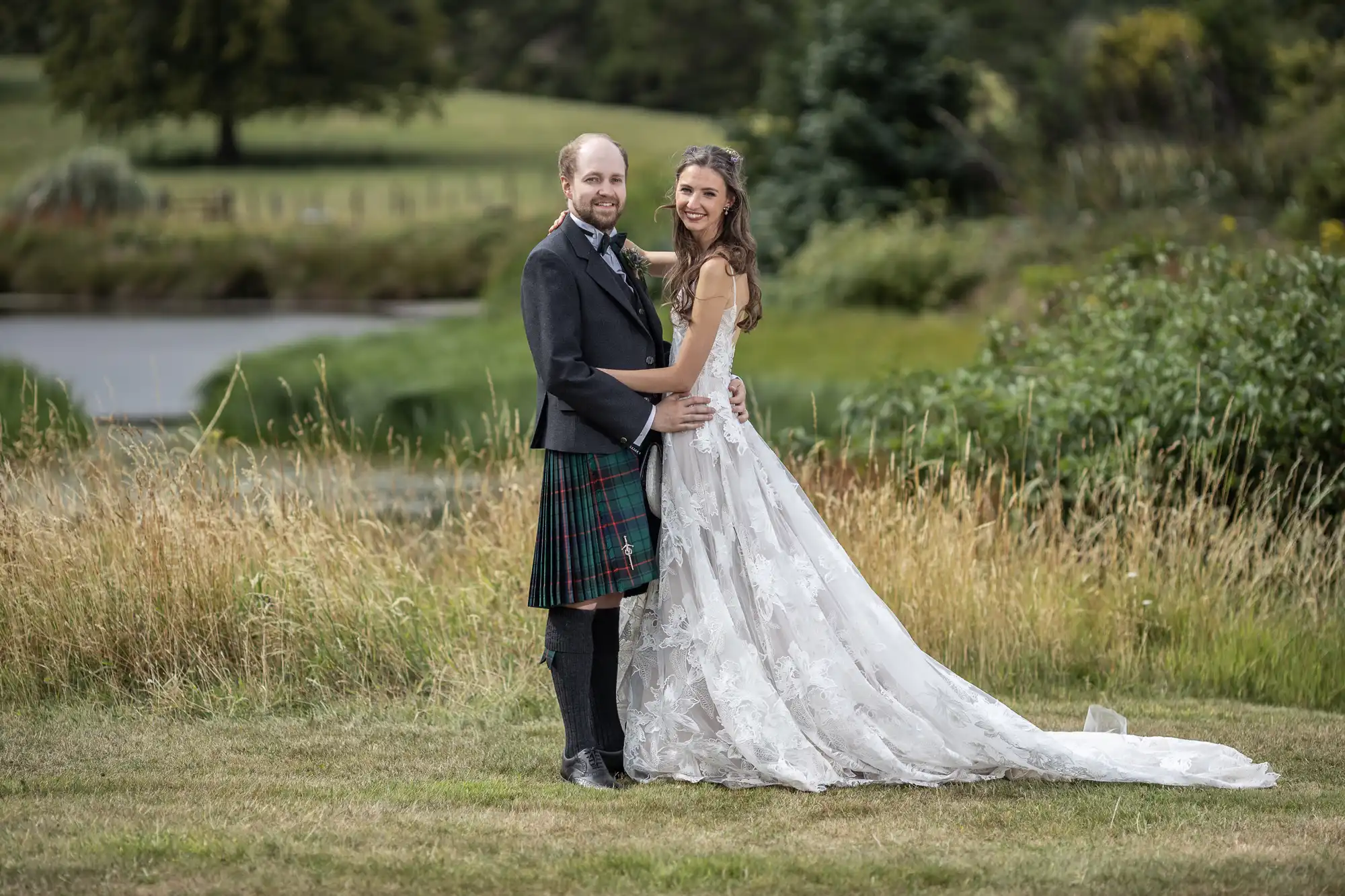 A couple standing outdoors in front of a pond and greenery, dressed in wedding attire. The groom is in a kilt, and the bride is in a white gown.