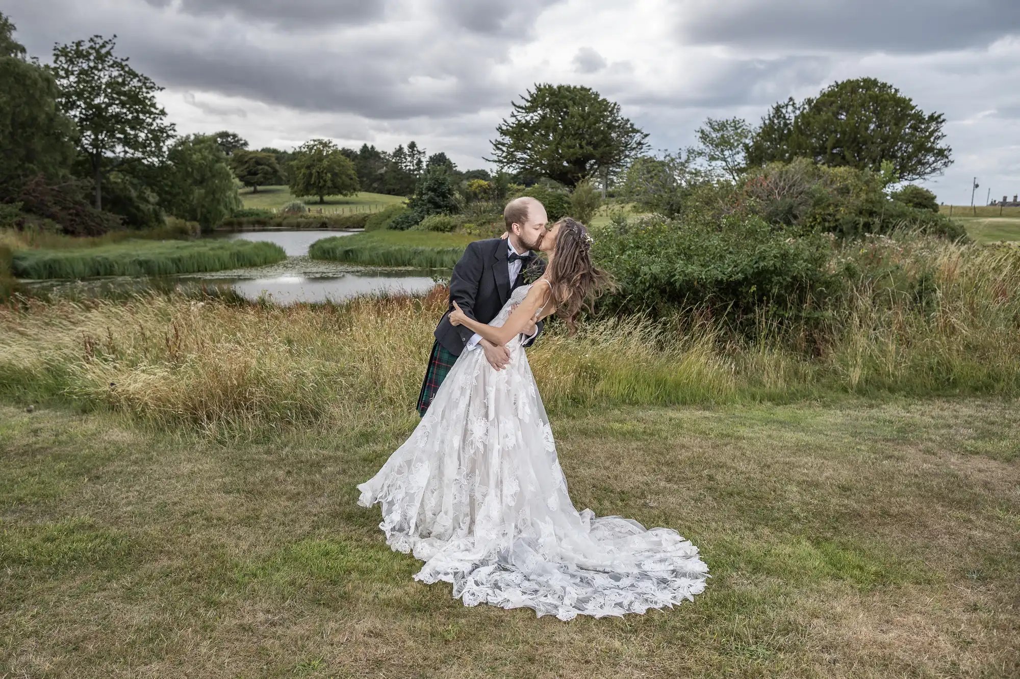 A couple in wedding attire shares a kiss outdoors near a pond, with the groom wearing a kilt. The scene includes lush greenery and a cloudy sky.